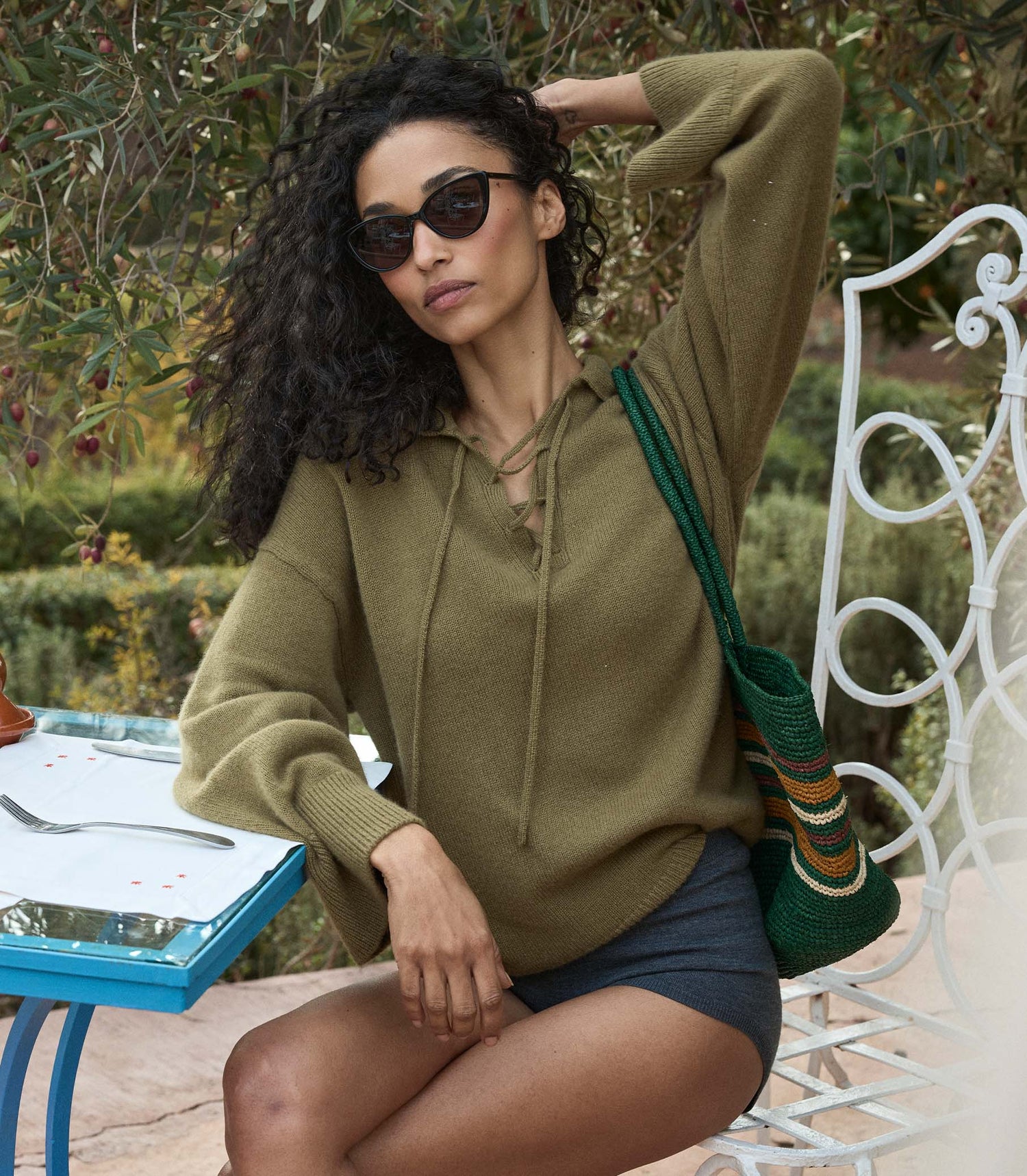 Woman in a green sweater and shorts sitting outdoors with a colorful bag over her shoulder.