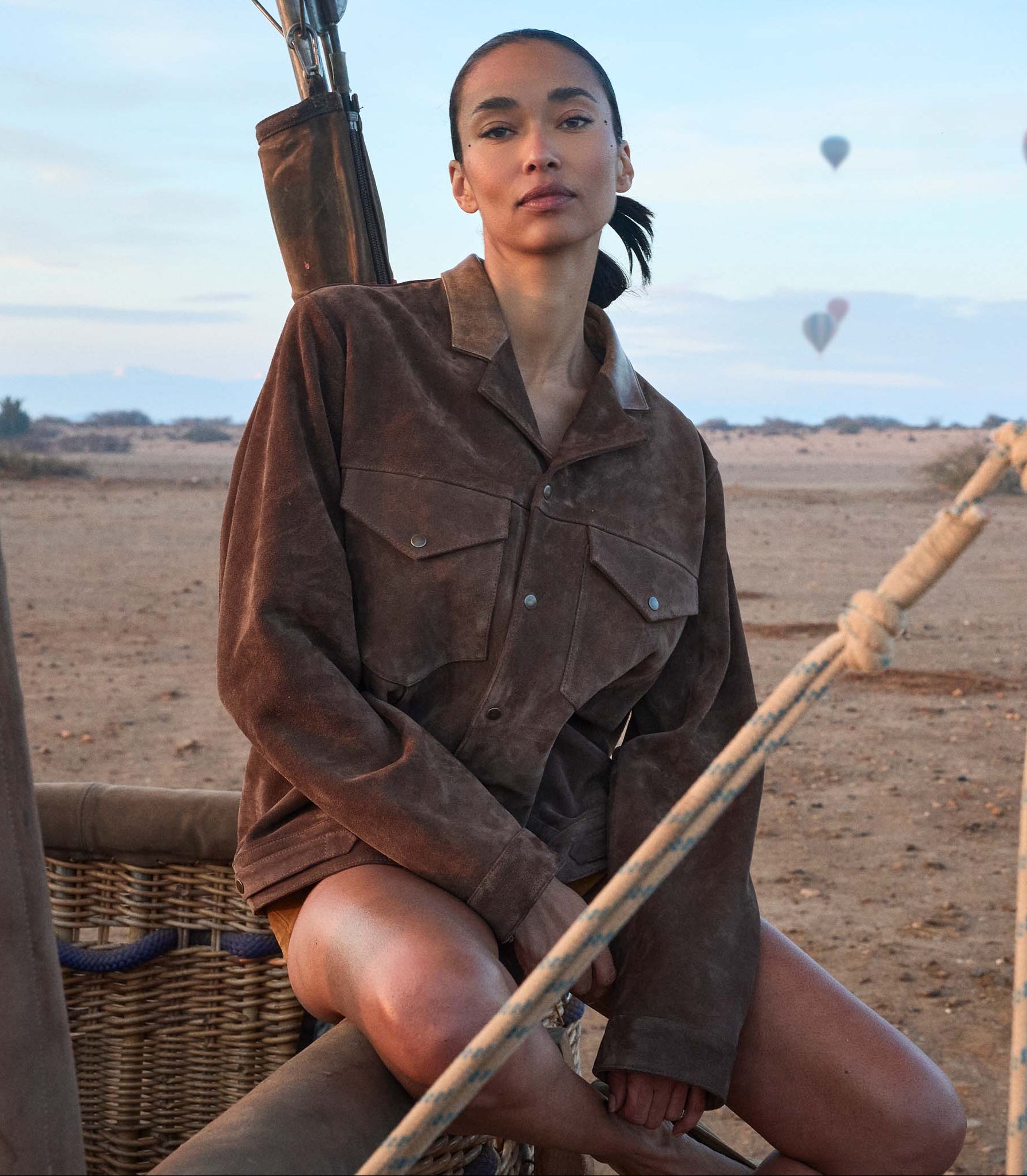 Person in a brown jacket sitting on a hot air balloon basket with balloons in the background 1
