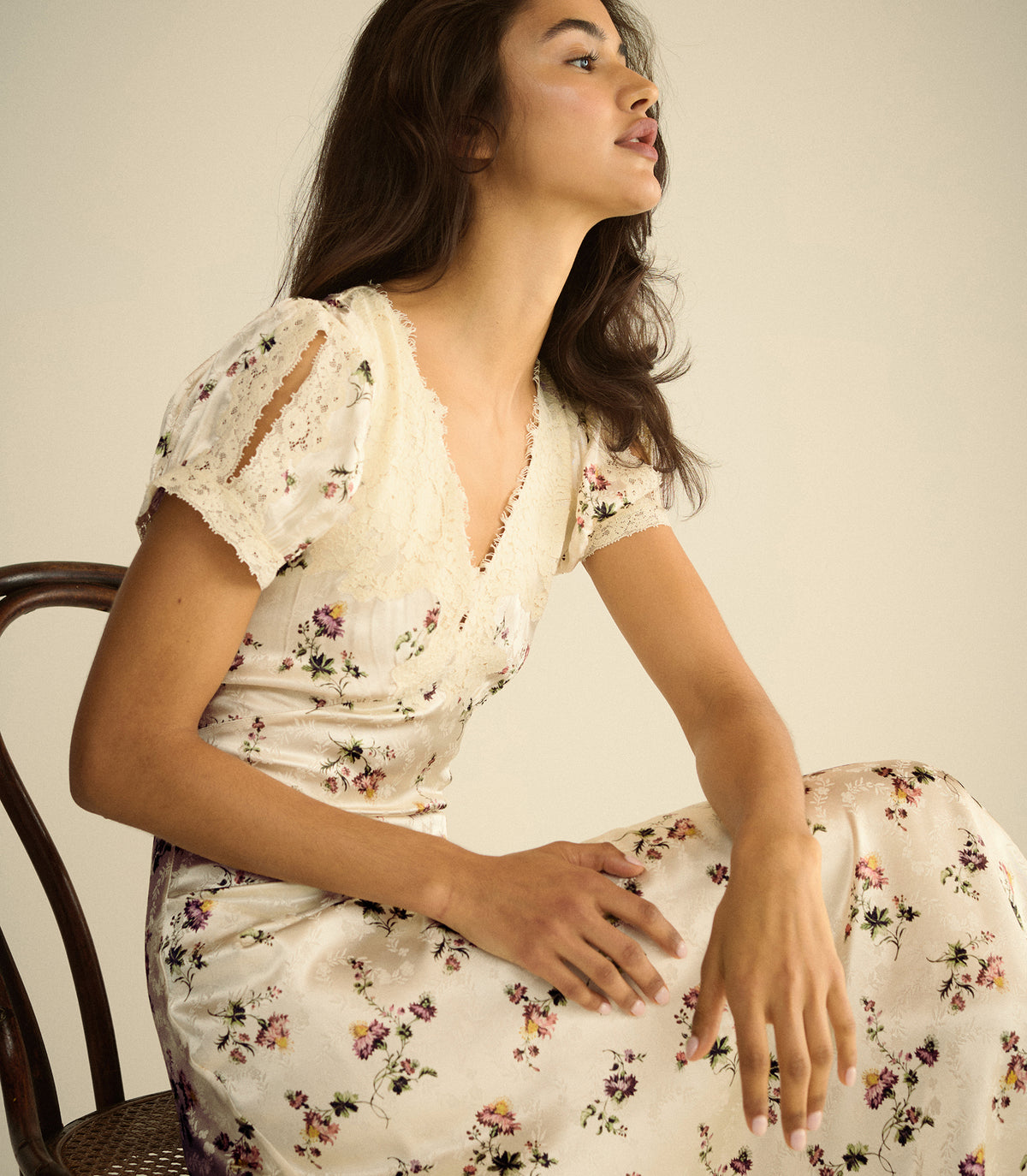 A woman wearing a cream-colored floral midi dress with lace puff sleeves sits on a wooden chair against a plain beige background.