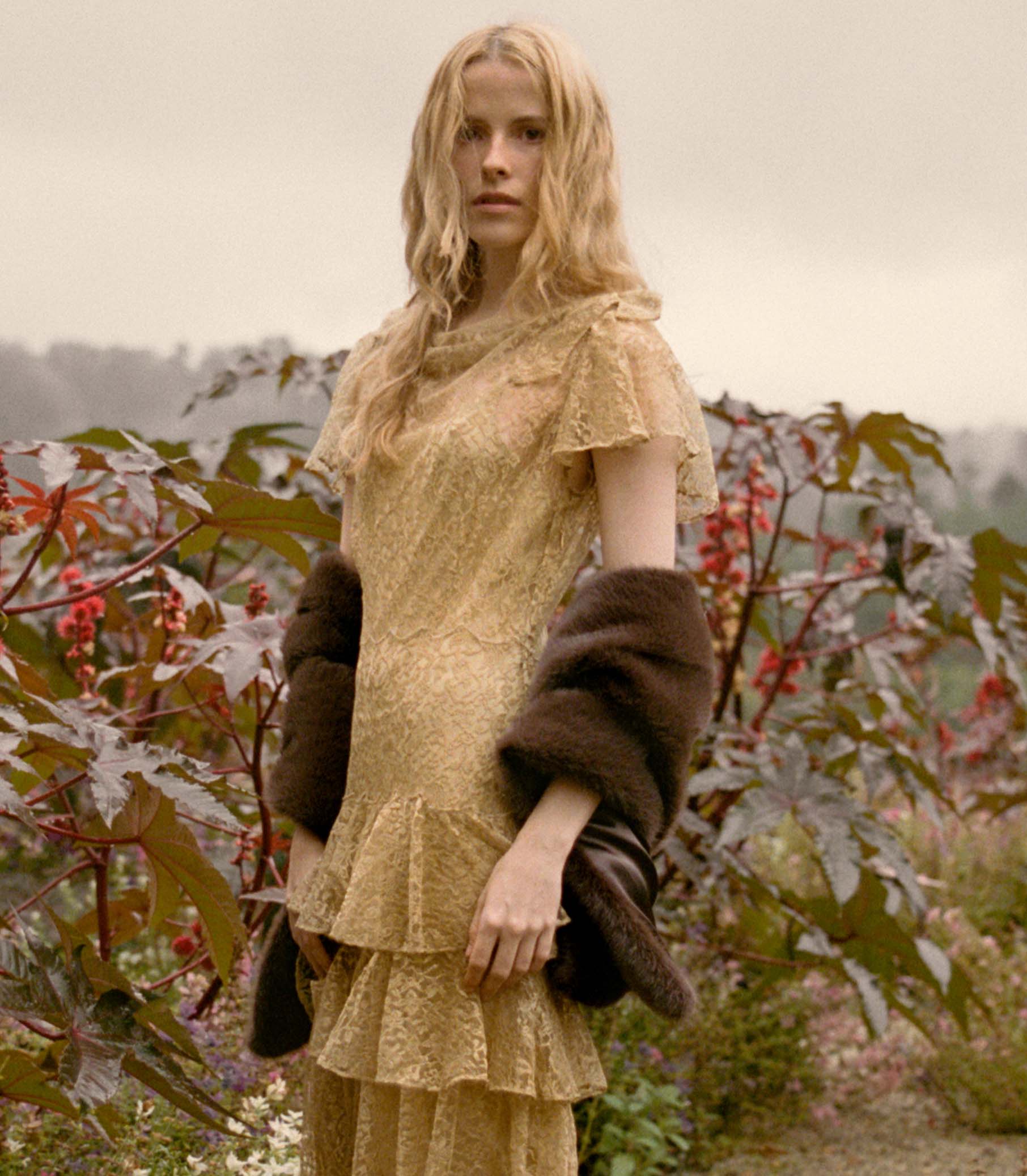 Woman in a gold lace dress holding a faux fur stole amidst autumn foliage. 1