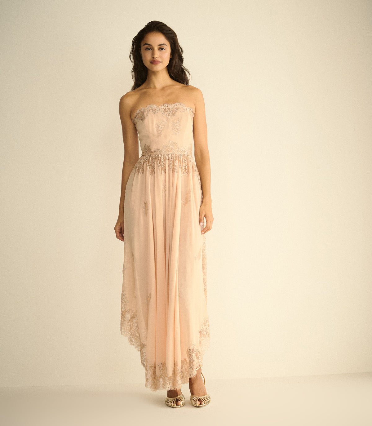 A woman modeling a strapless peach maxi dress with lace detailing on the bodice and hem, paired with metallic gold high-heeled sandals, set against a plain cream studio background. 2
