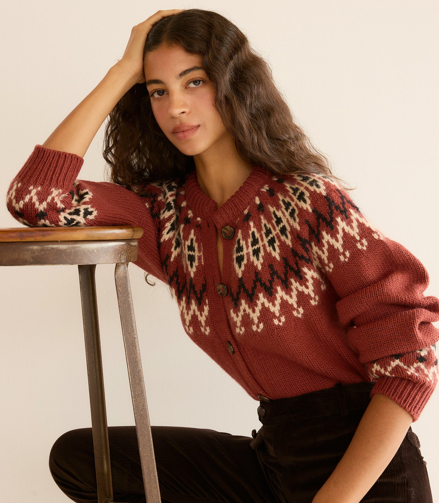 Woman wearing a patterned sweater sitting on a stool against a plain background