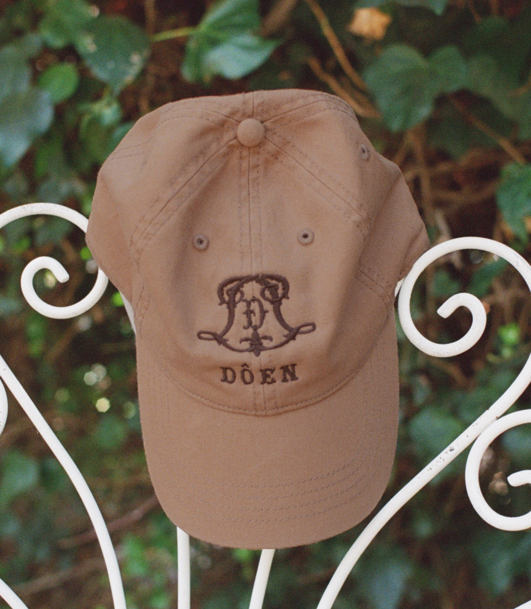 A tan baseball cap with a dark brown embroidered monogram and the word 'DÔEN' on the front, displayed on a white wrought iron garden chair with green foliage in the background. 1
