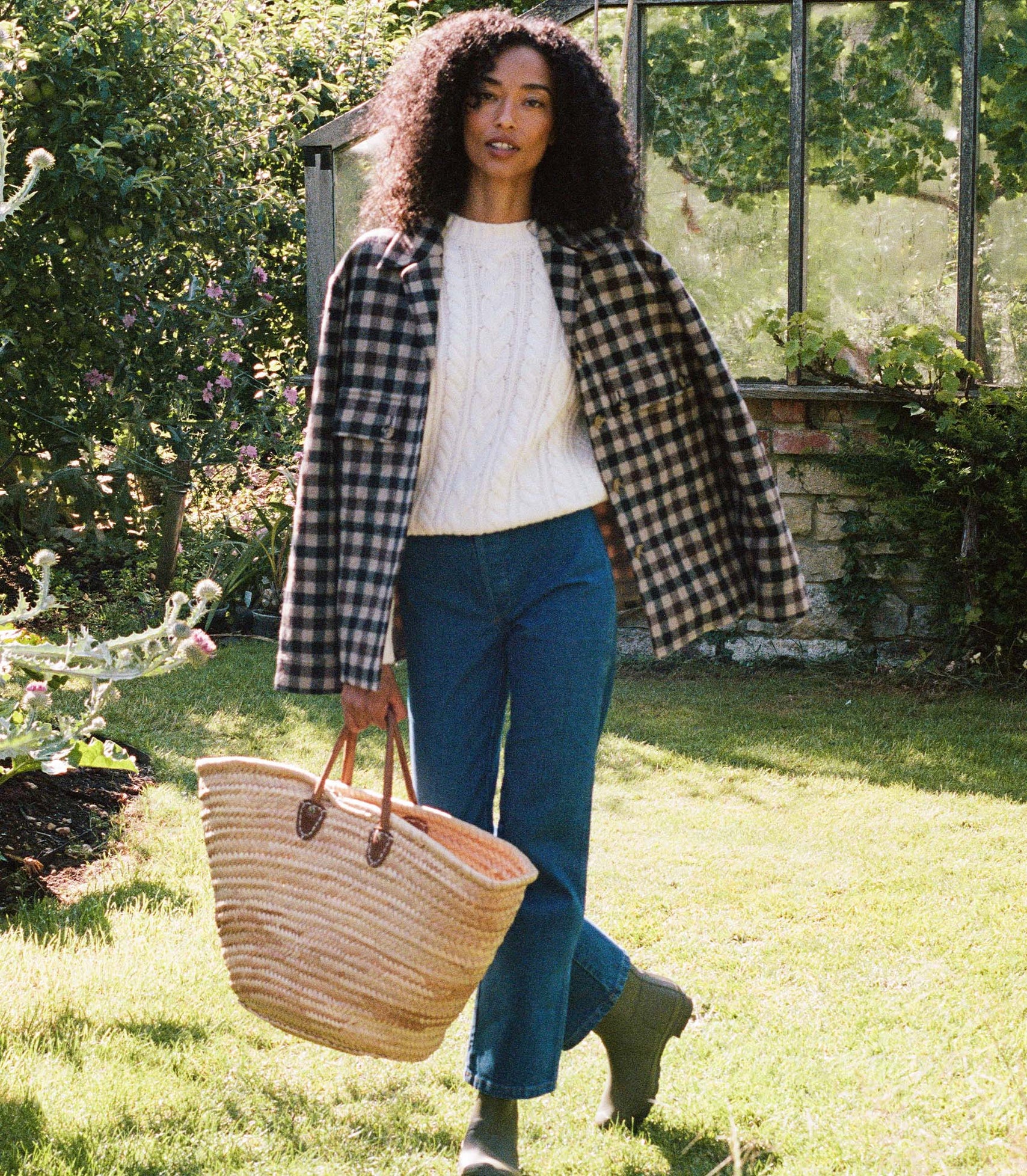 Person standing in a garden and wearing a checkered plaid jacket, white sweater, and jeans.