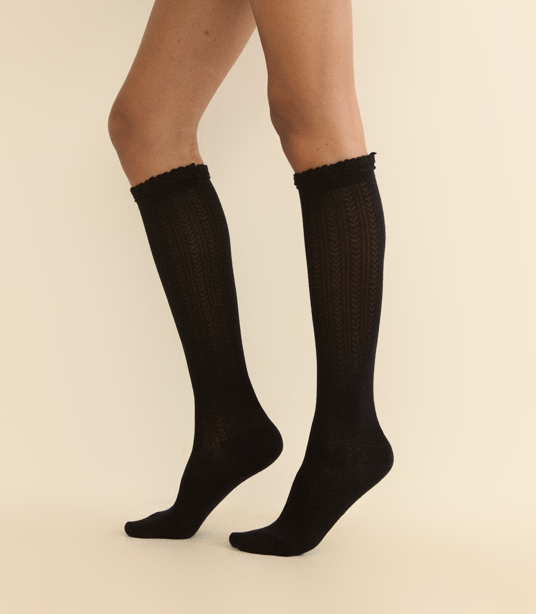 Black knee-high cable-knit socks worn on a model's legs, shown against a plain beige background. 1
