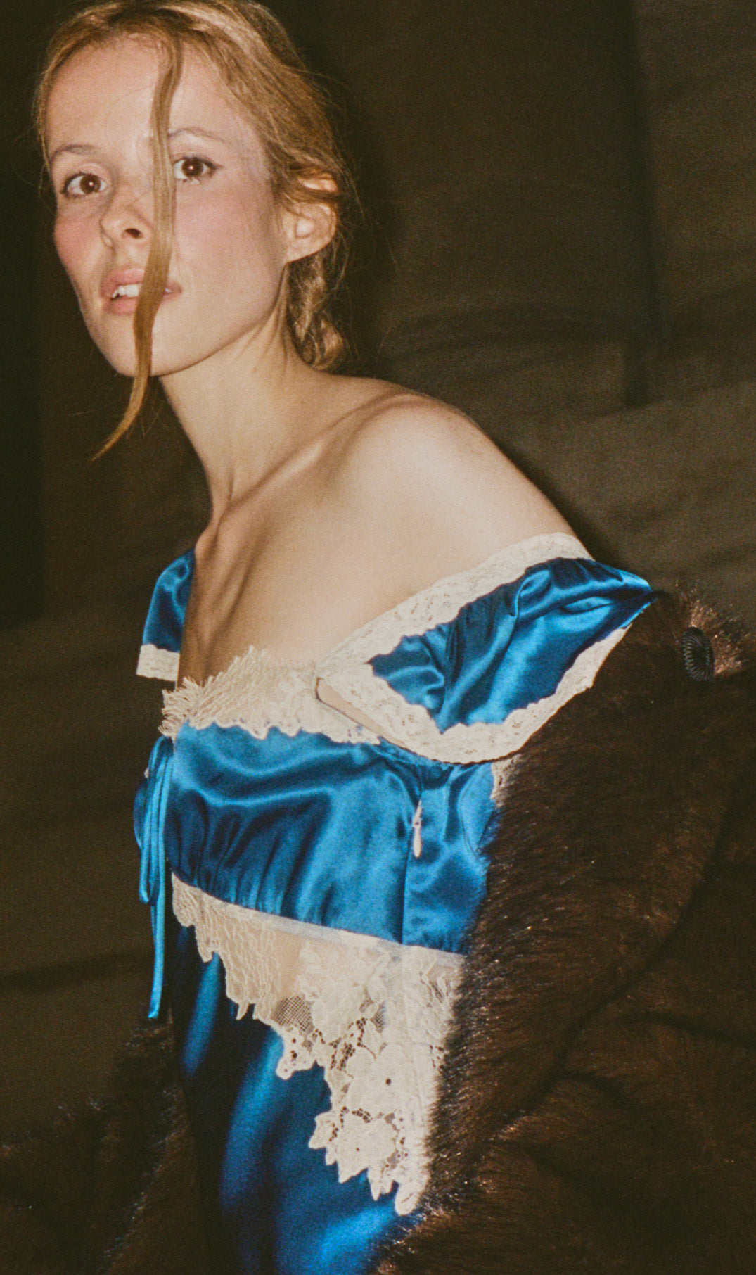 A woman wearing a shiny royal blue satin off-the-shoulder dress with cream lace trim and a dark brown fur collar, photographed in dim warm-toned lighting against a dark indoor background.