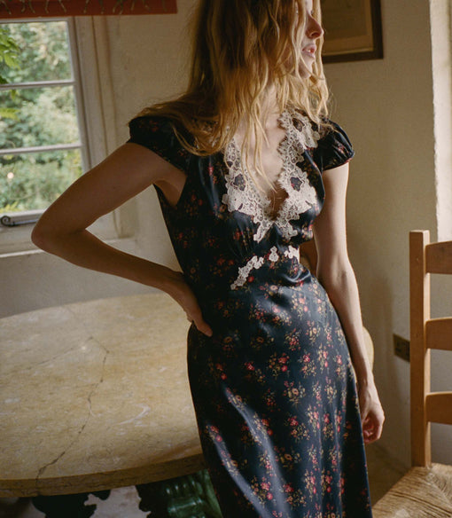 Woman wearing a black floral dress with lace details in a room with a window and chair.