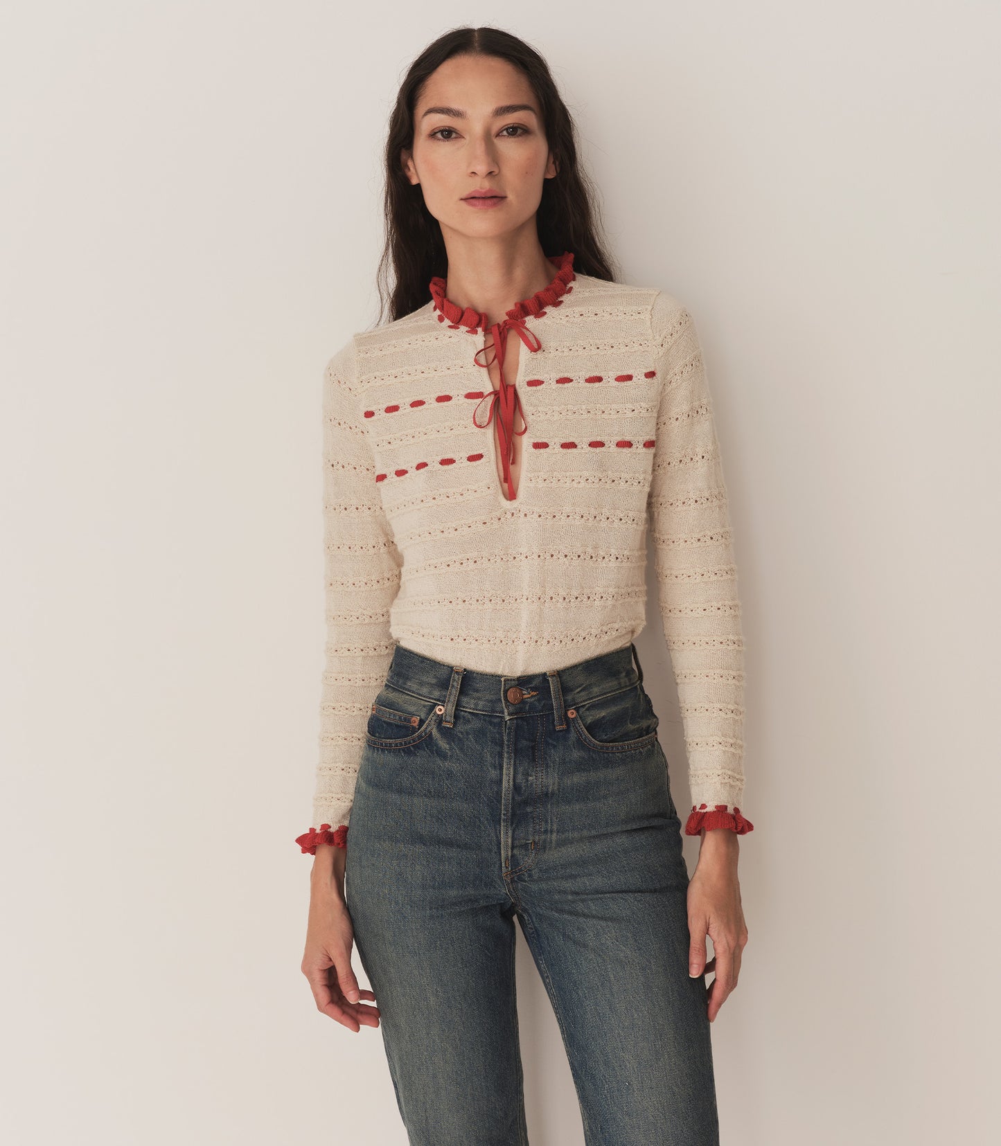 A woman wearing jeans and the Cordelie Top, in a cream and red baby alpaca-silk blend and silk grosgrain ribbons that tie at the neck.