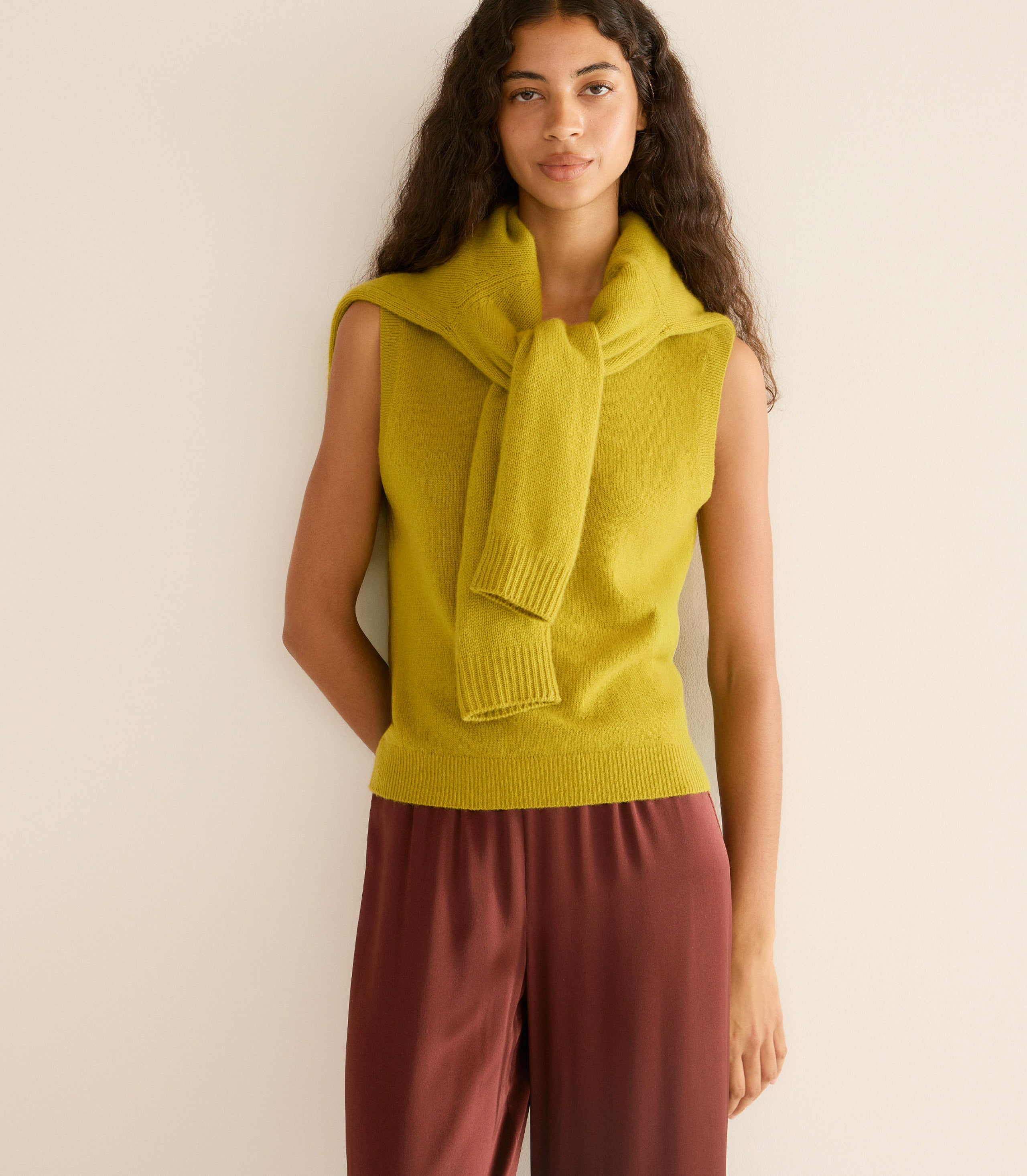 Woman wearing a mustard yellow sleeveless sweater with a matching scarf against a beige background