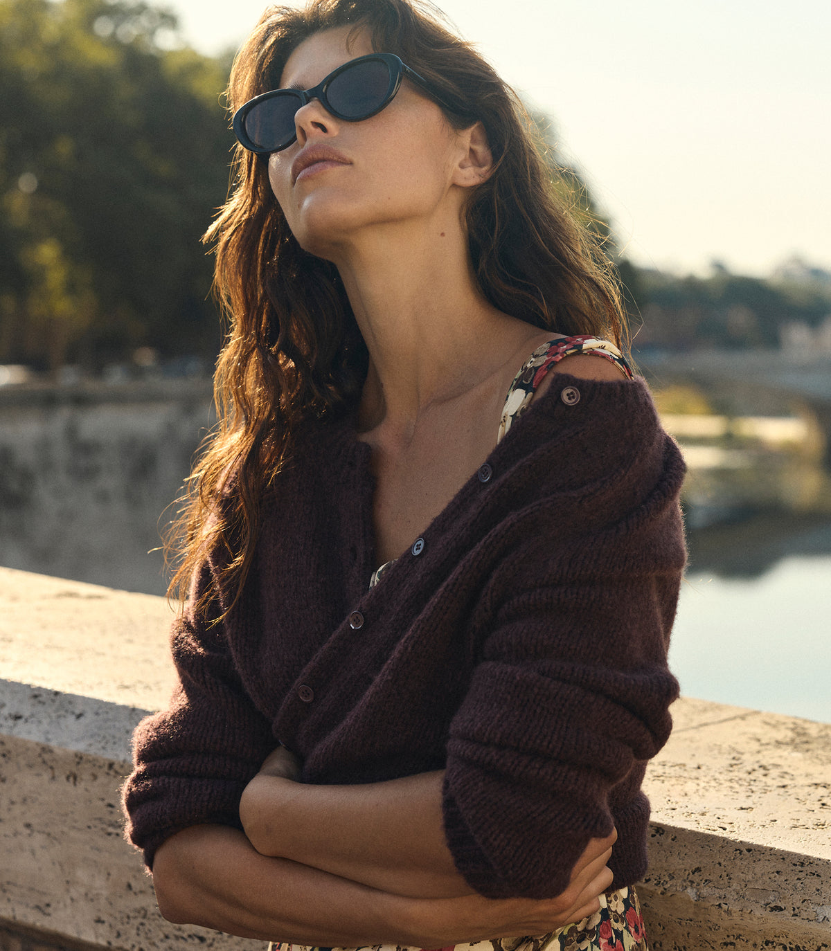 A woman wearing dark sunglasses, a dark brown knit cardigan, and a floral dress, leaning against a stone wall with a river and trees in the background. 1
