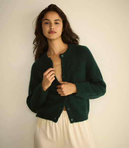 A woman wearing a dark forest green knit cardigan with buttons, paired with a cream-colored skirt, posing against a plain beige wall.