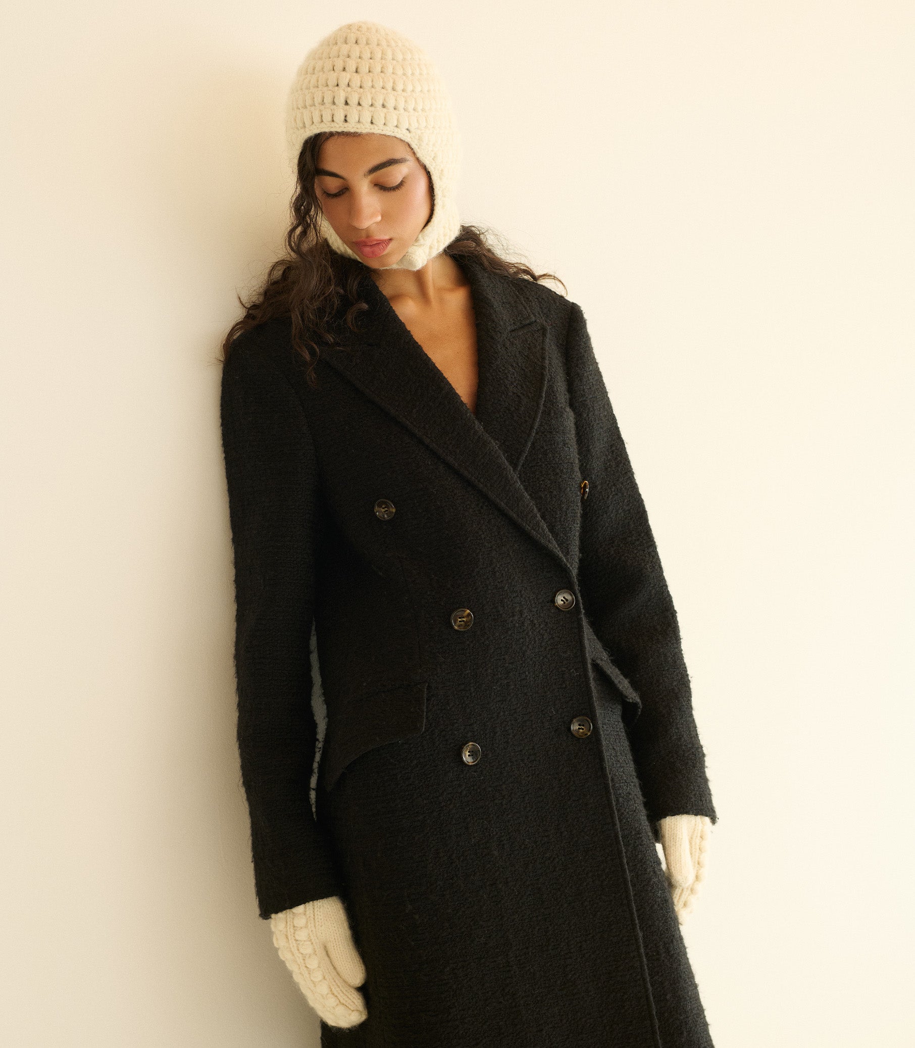 Woman wearing a black coat, white beanie, and gloves against a plain background