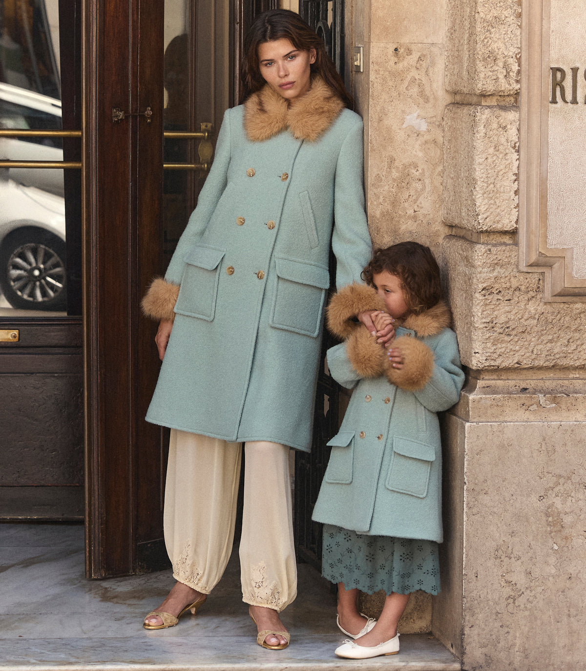 A woman and a young girl wearing matching sage green wool coats with brown fur-trimmed collars and gold buttons, standing on a stone building entrance.