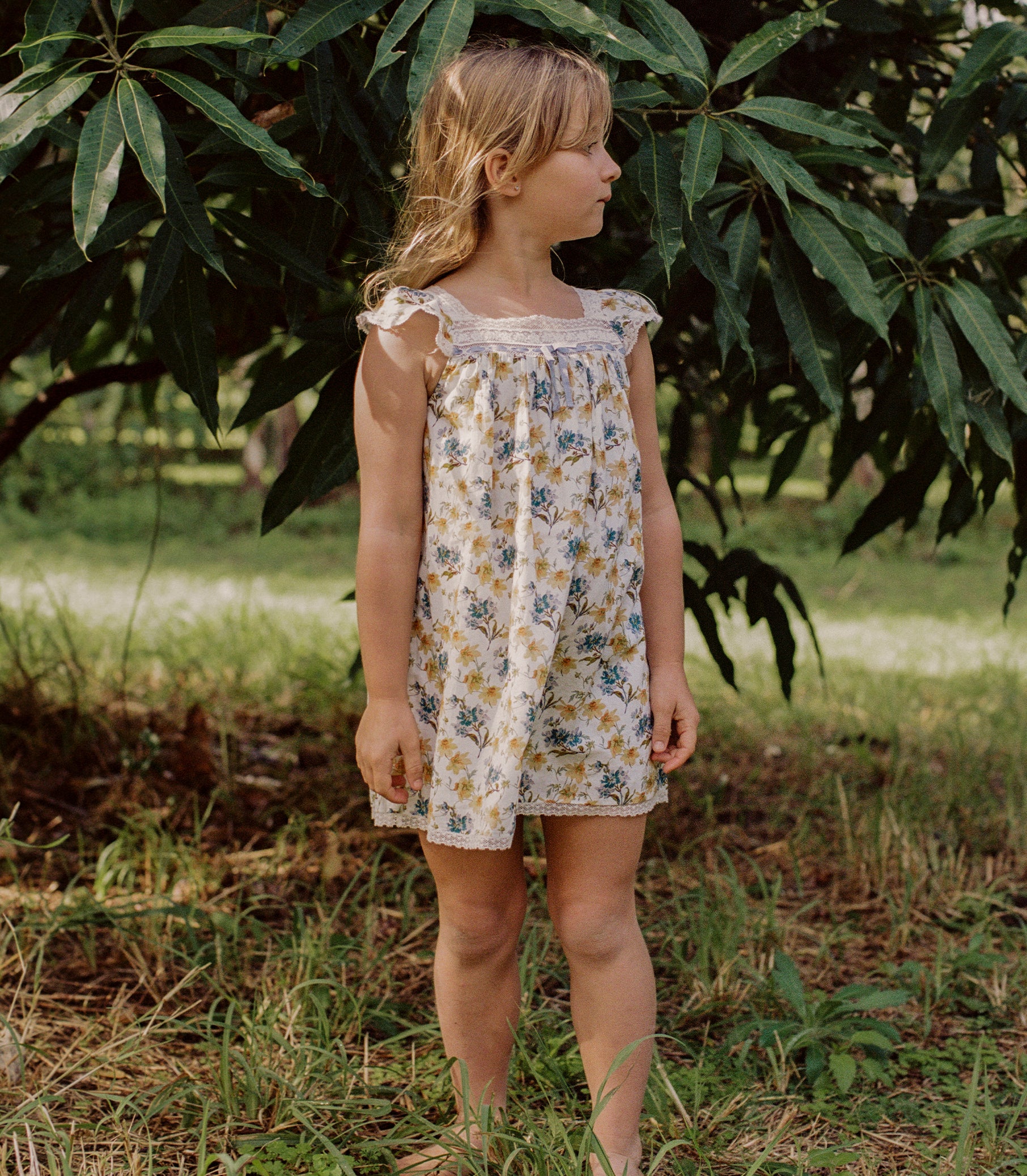 A young girl wearing a sleeveless cream-colored dress with a blue and yellow floral print, standing outdoors in a grassy area under leafy green trees. 1

