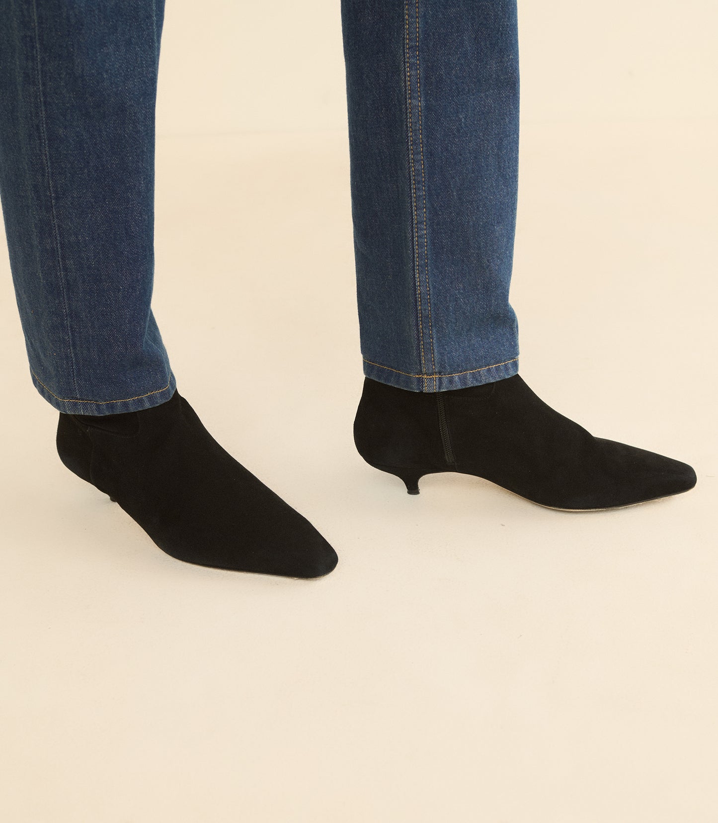 Black pointed-toe ankle boots with kitten heels, shown on a model wearing blue jeans against a plain beige background.