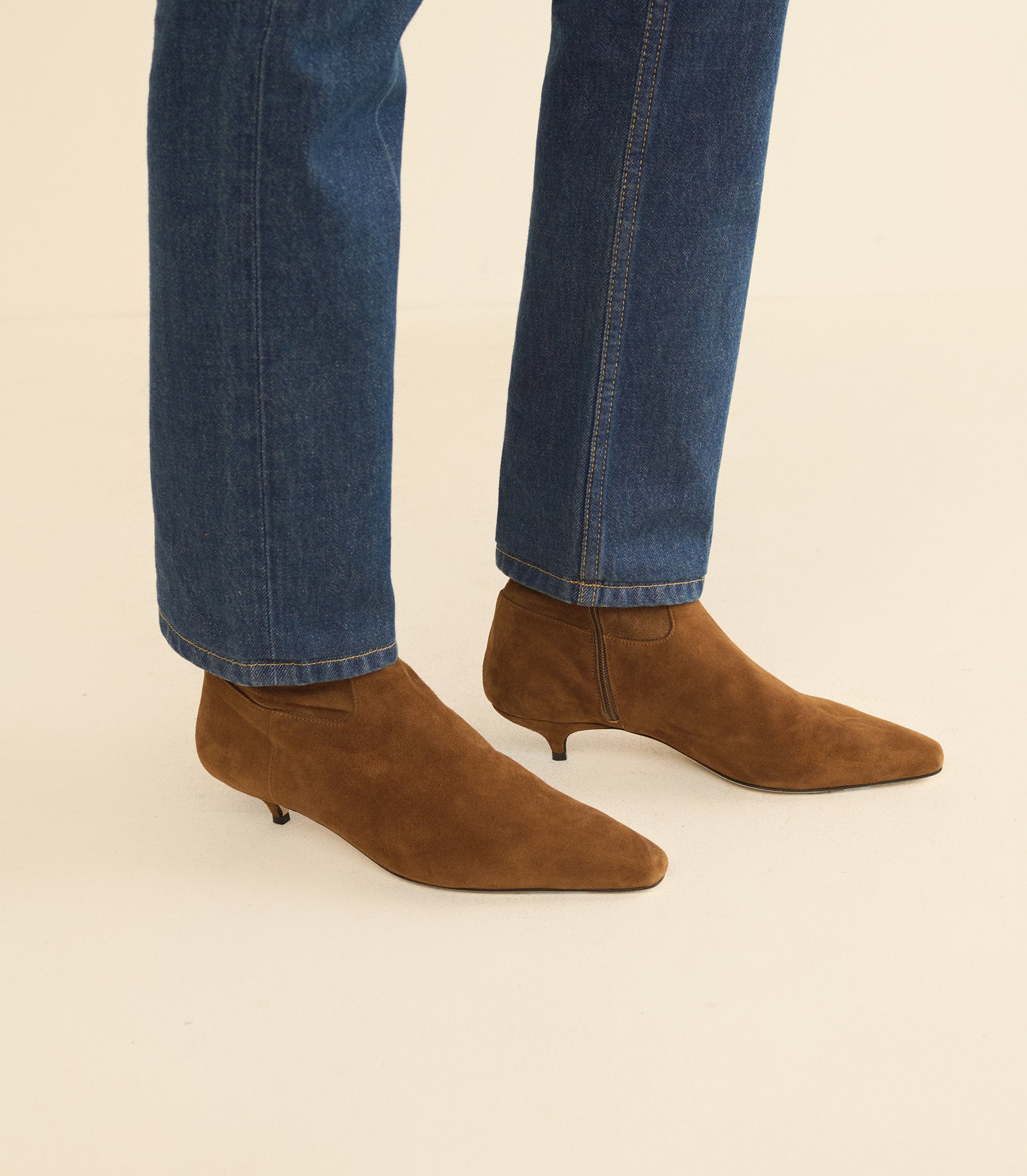 A person wearing pointed-toe brown suede ankle boots with kitten heels, paired with blue jeans, shown from the calves down against a plain light beige background.
