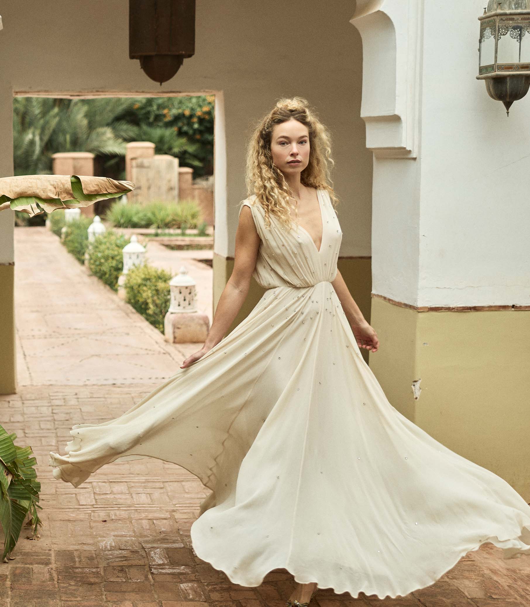 Woman in a flowing white dress standing in a sunlit outdoor setting with plants and a building. 1
