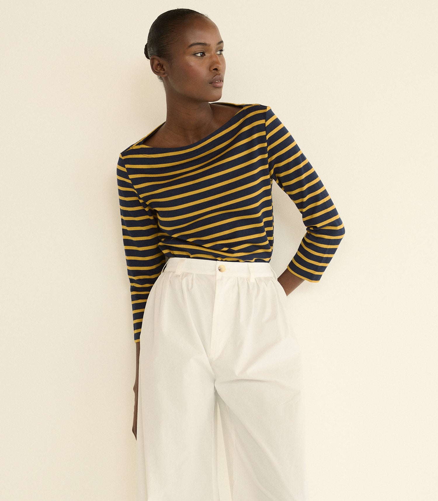 A model wearing a navy and mustard yellow striped long-sleeve boat-neck top paired with white high-waisted trousers, standing against a plain cream-colored wall.