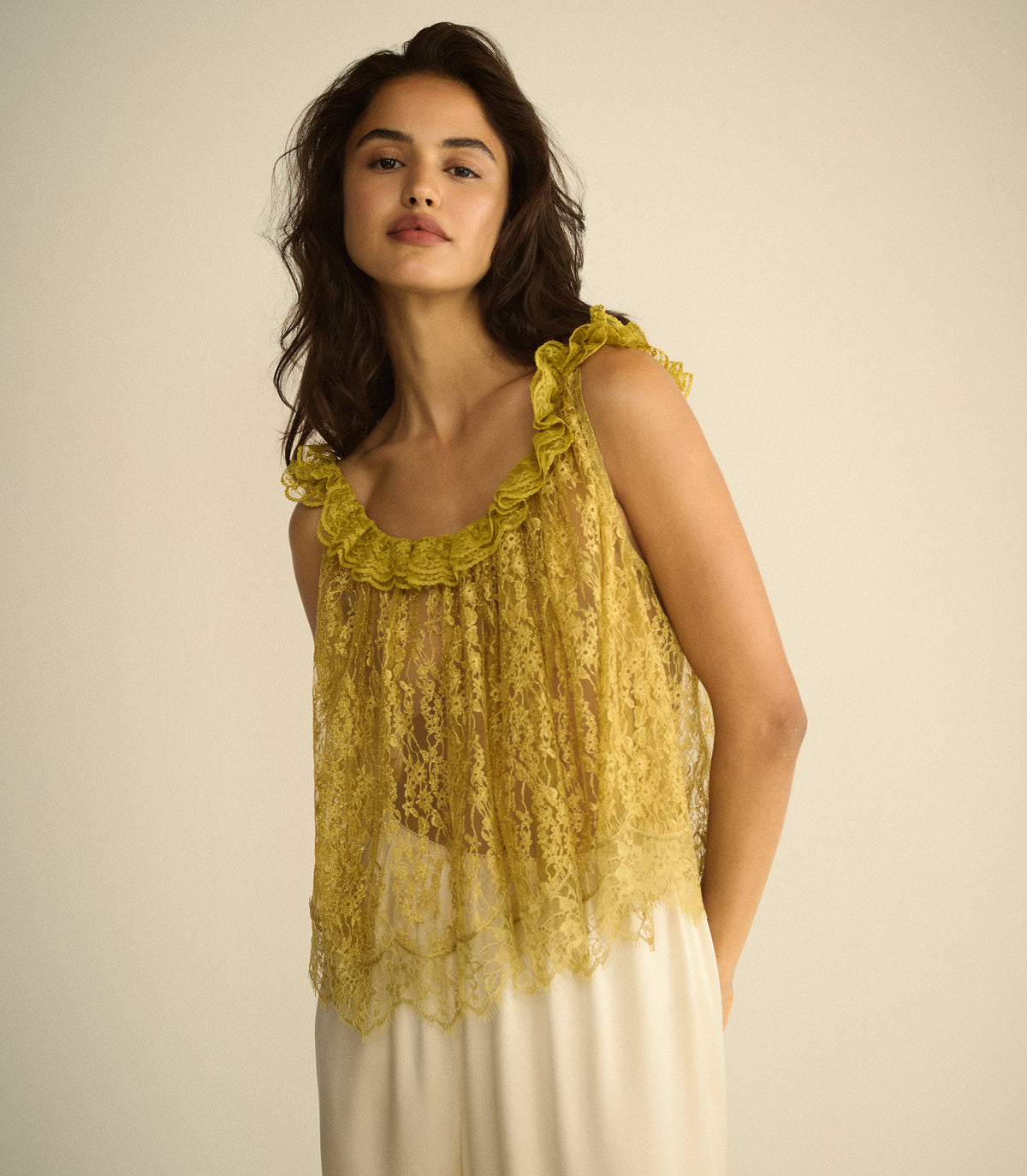 A woman wearing a yellow lace sleeveless top with ruffled trim and a white skirt, standing against a plain beige background.