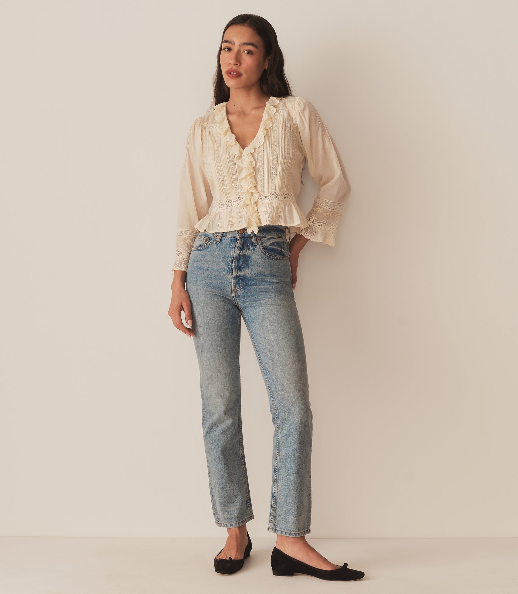 A woman wearing the Eldoris Top in Alabaster, made in organic cotton voile and featuring a plunging V-neckline framed with cascading lace-trimmed ruffles that extend down the bodice and alternating panels of inset lace, clustered pintucks, and intricate embroidery at the yoke and back.