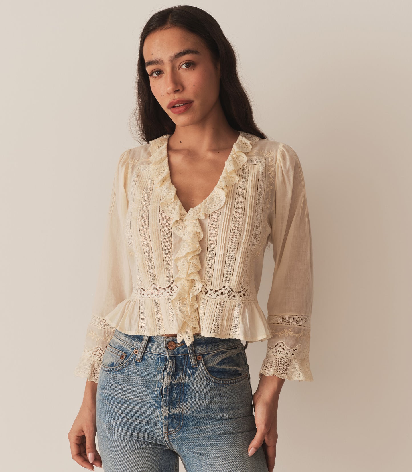 A woman wearing the Eldoris Top in Alabaster, made in organic cotton voile and featuring a plunging V-neckline framed with cascading lace-trimmed ruffles that extend down the bodice and alternating panels of inset lace, clustered pintucks, and intricate embroidery at the yoke and back.