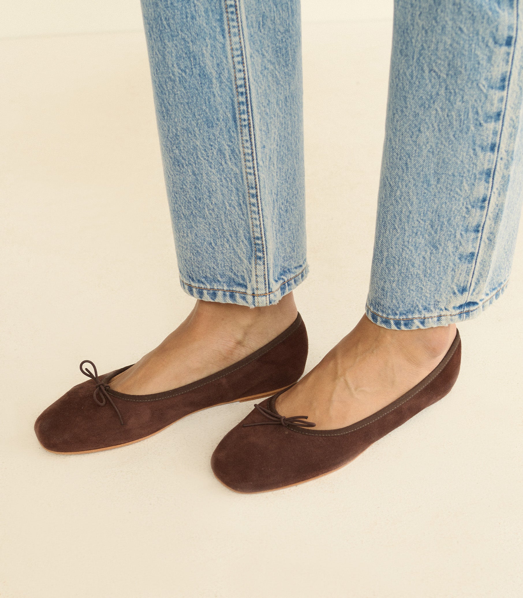 A person wearing dark brown suede ballet flats with small bow details, paired with light-wash blue jeans, shown from the calves down against a plain light beige background. 1
