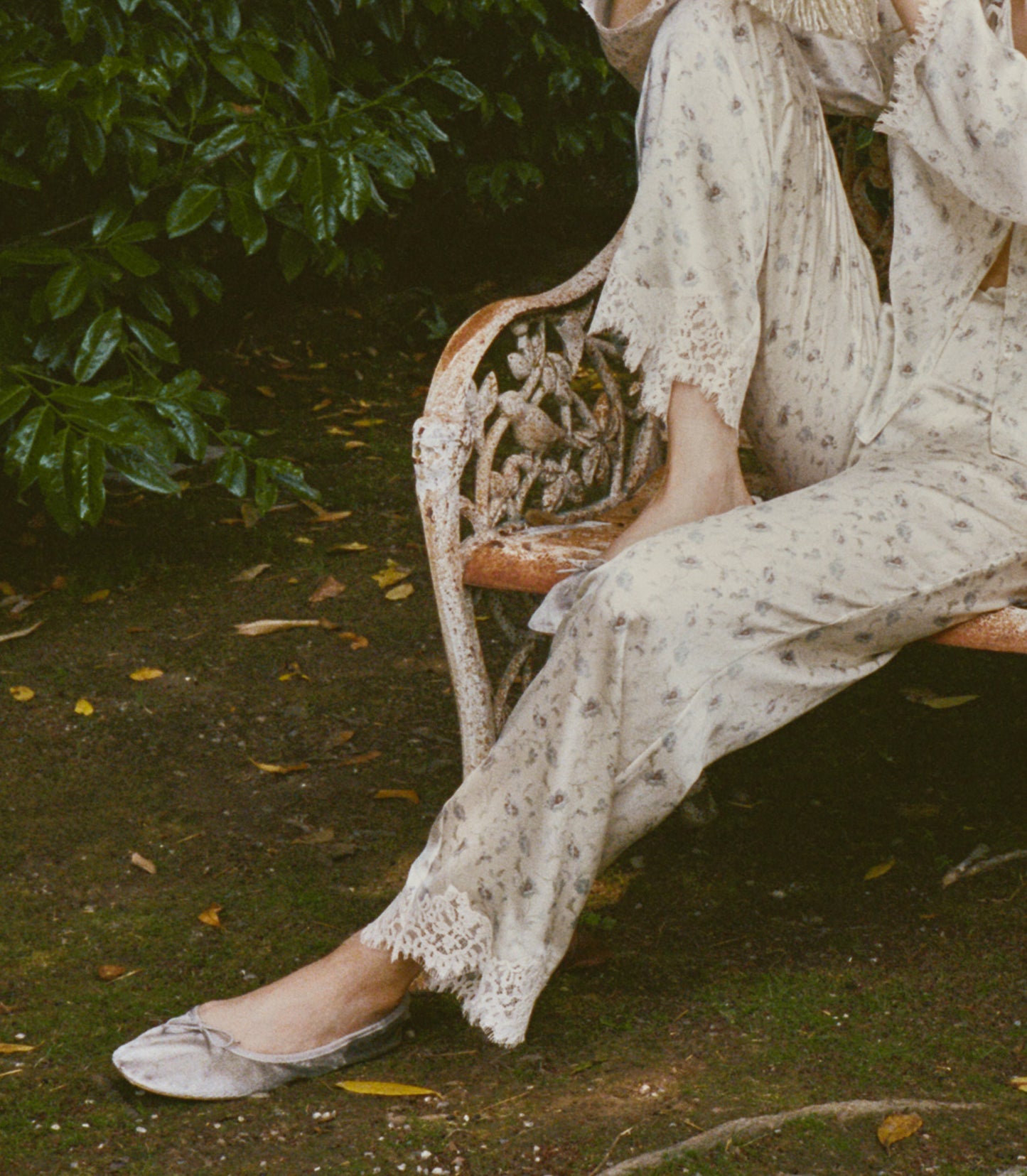 A person seated on an outdoor garden bench wearing a loose cream-colored silk pants with lace trim and light flats, surrounded by green foliage and a dirt ground.
