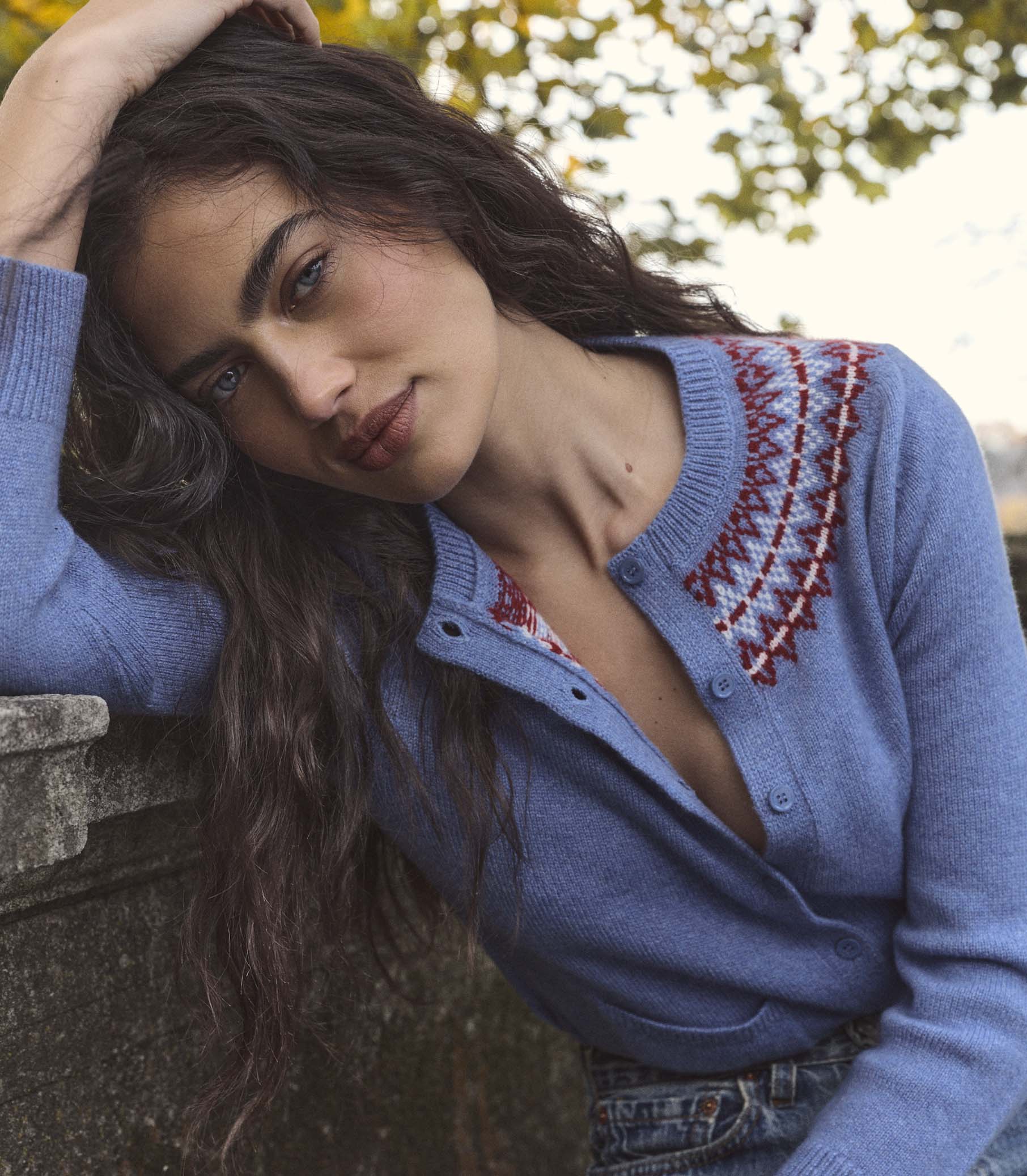 Woman wearing a blue cardigan with intricate patterns, sitting outdoors.