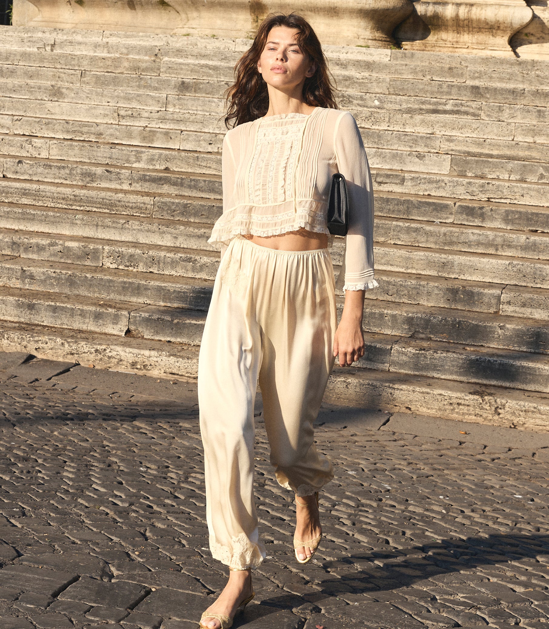 A woman walking outdoors on stone steps wearing a cream-colored two-piece outfit consisting of a sheer long-sleeve crop top with lace detailing and matching loose trousers. She is carrying a small black shoulder bag. 1
