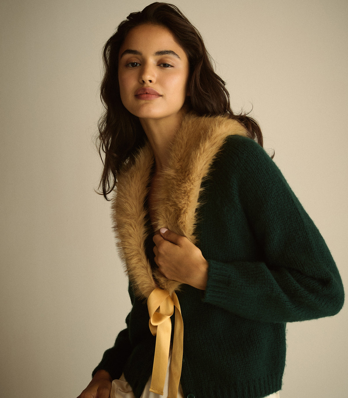 A woman wearing a dark green knit sweater with a tan faux-fur collar and a matching tan ribbon tie, photographed against a plain light beige studio background. 1
