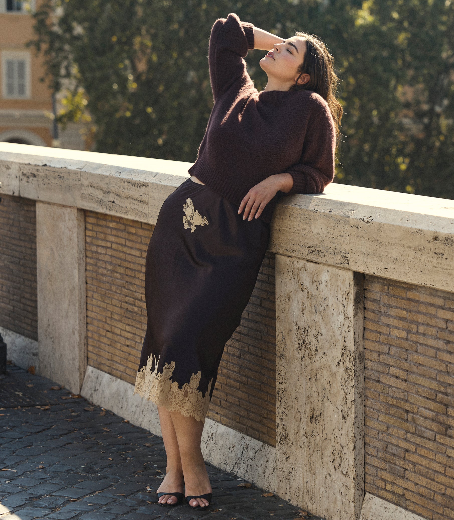 A woman leans against a stone bridge railing outdoors, wearing a dark brown knit sweater and matching midi skirt with beige floral embroidery and a beige lace hem. 1
