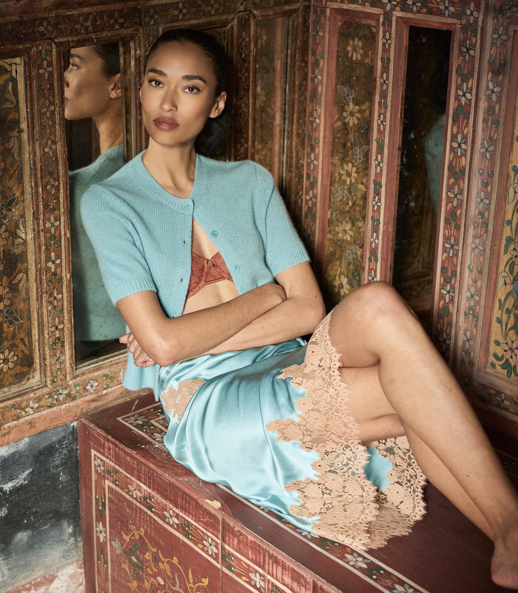 Woman in an aquamarine cardigan and lace silk skirt sitting on an ornate wooden bench.