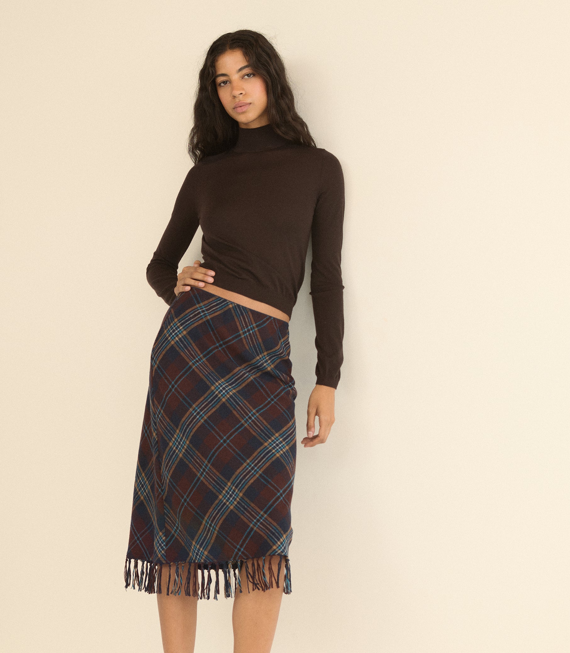 A woman modeling a dark brown long-sleeve mock neck crop top and a dark red and blue tartan plaid wrap skirt with fringe, standing against a plain beige background.