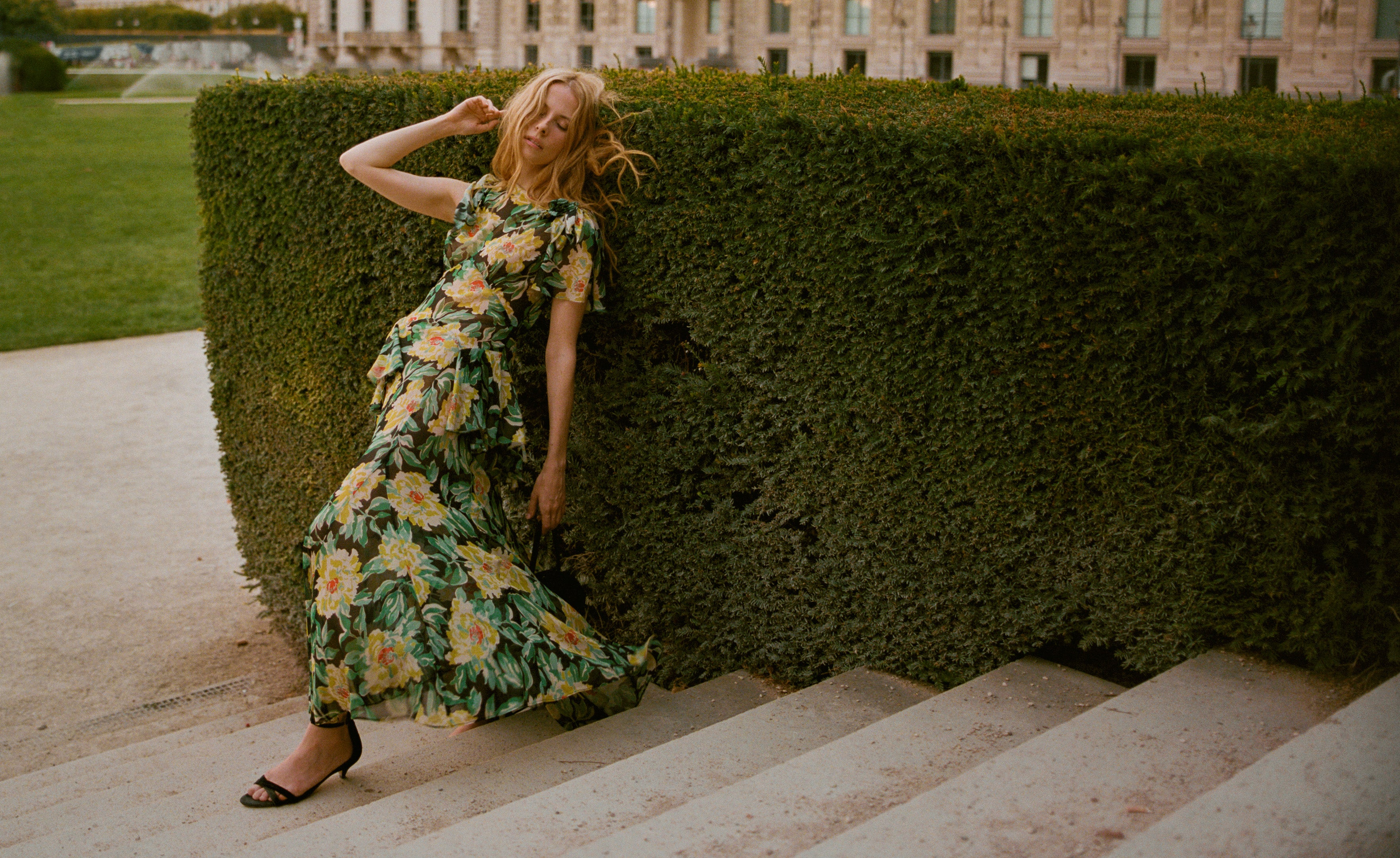 A woman in a multicolor floral dress leaning against shrubbery on steps.