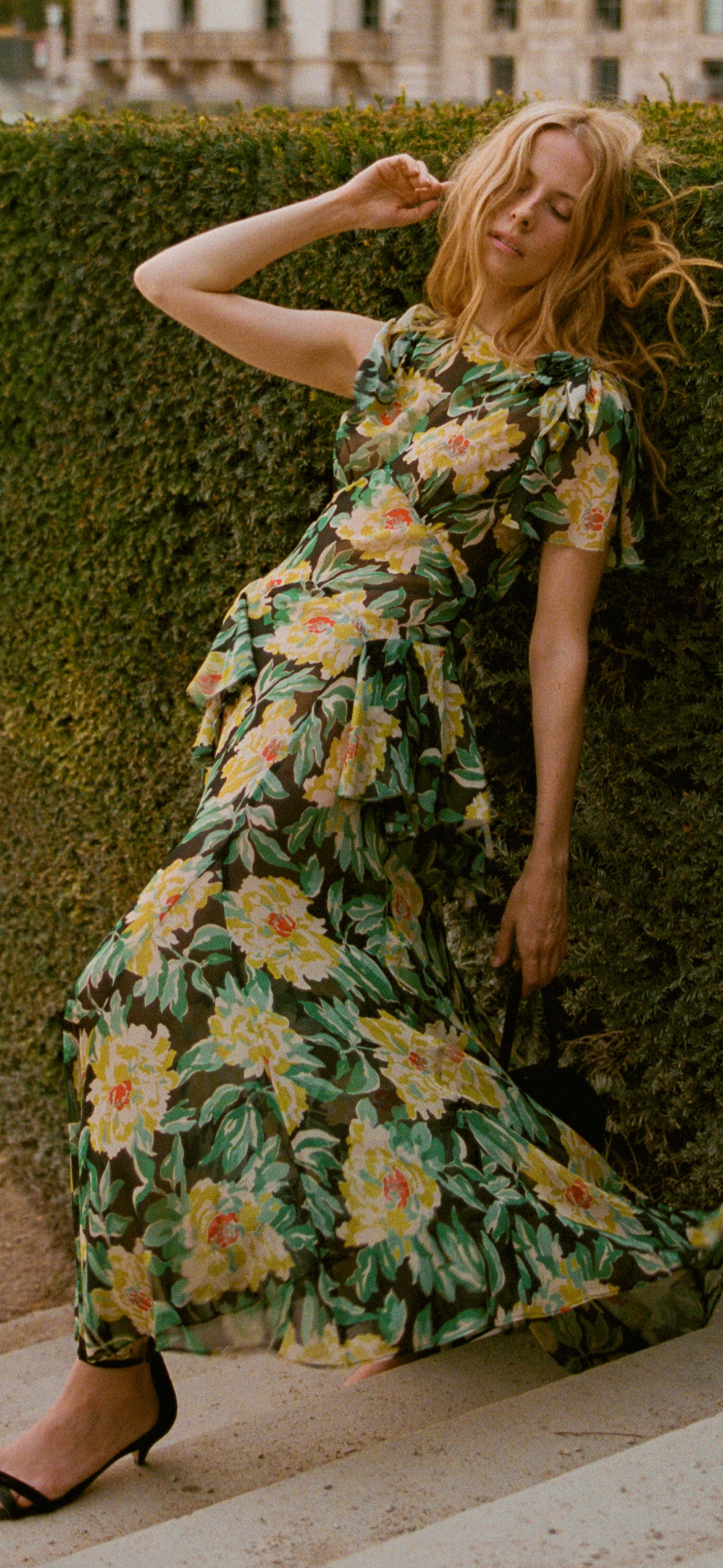 A woman in a multicolor floral dress leaning against shrubbery on steps.
