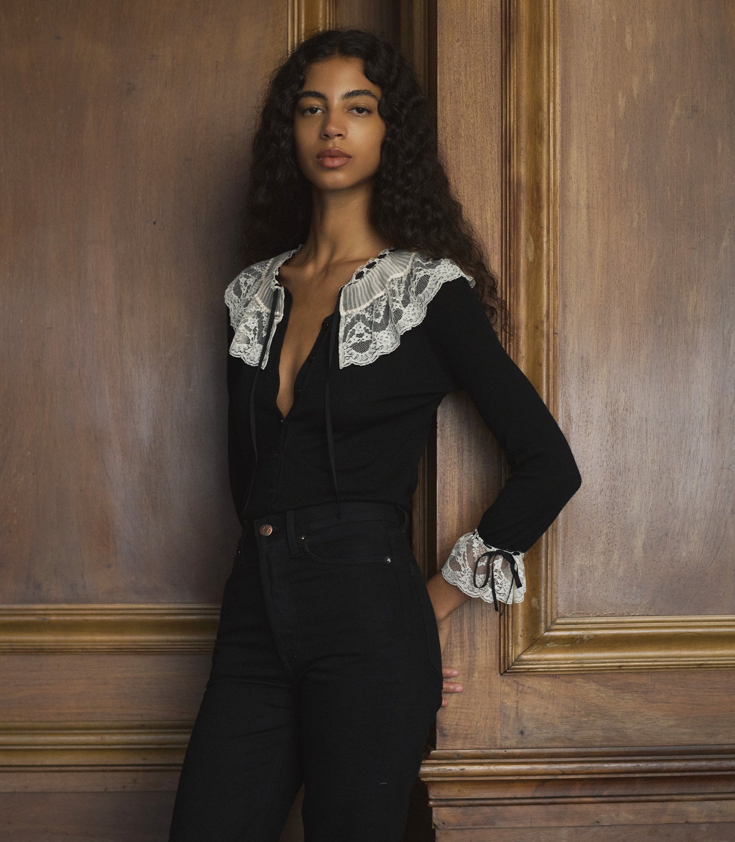 Woman wearing a black cardigan with with white lace details against a wooden wall.