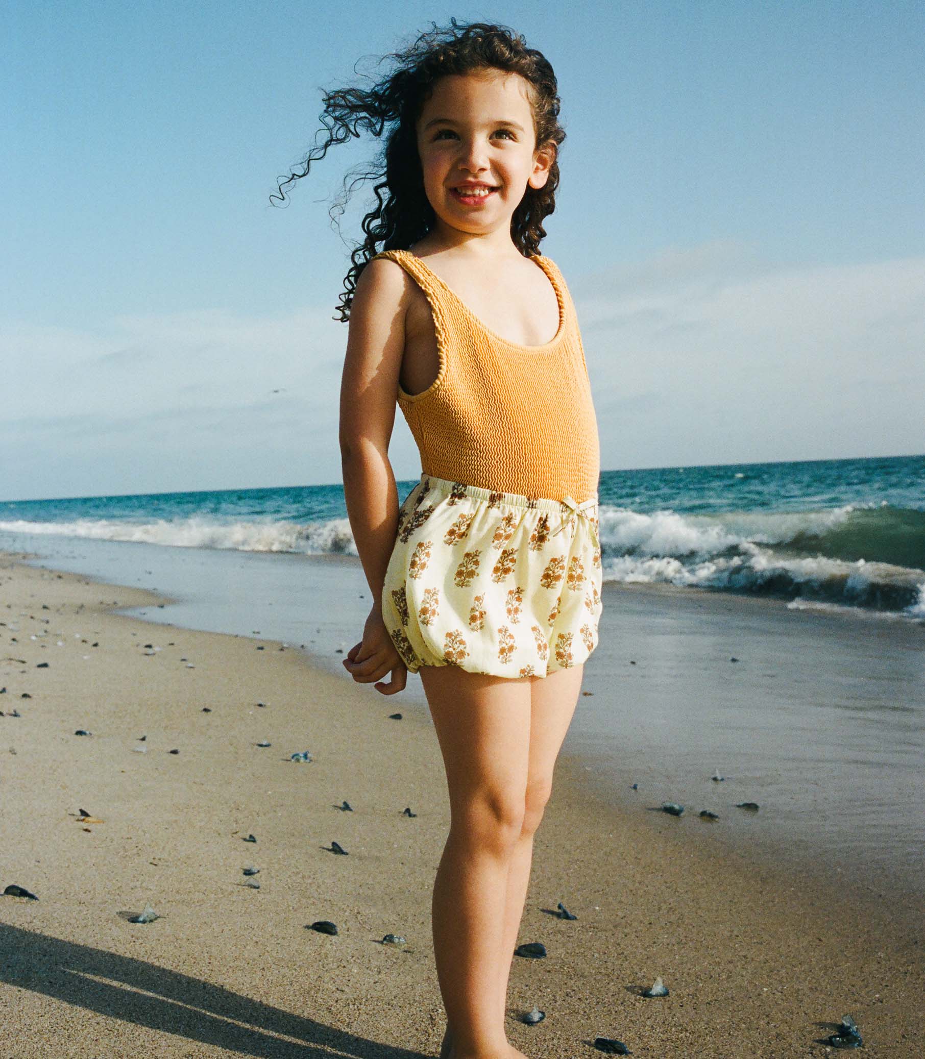 Young girl in a yellow top and floral shorts standing on a beach with ocean waves in the background. 1
