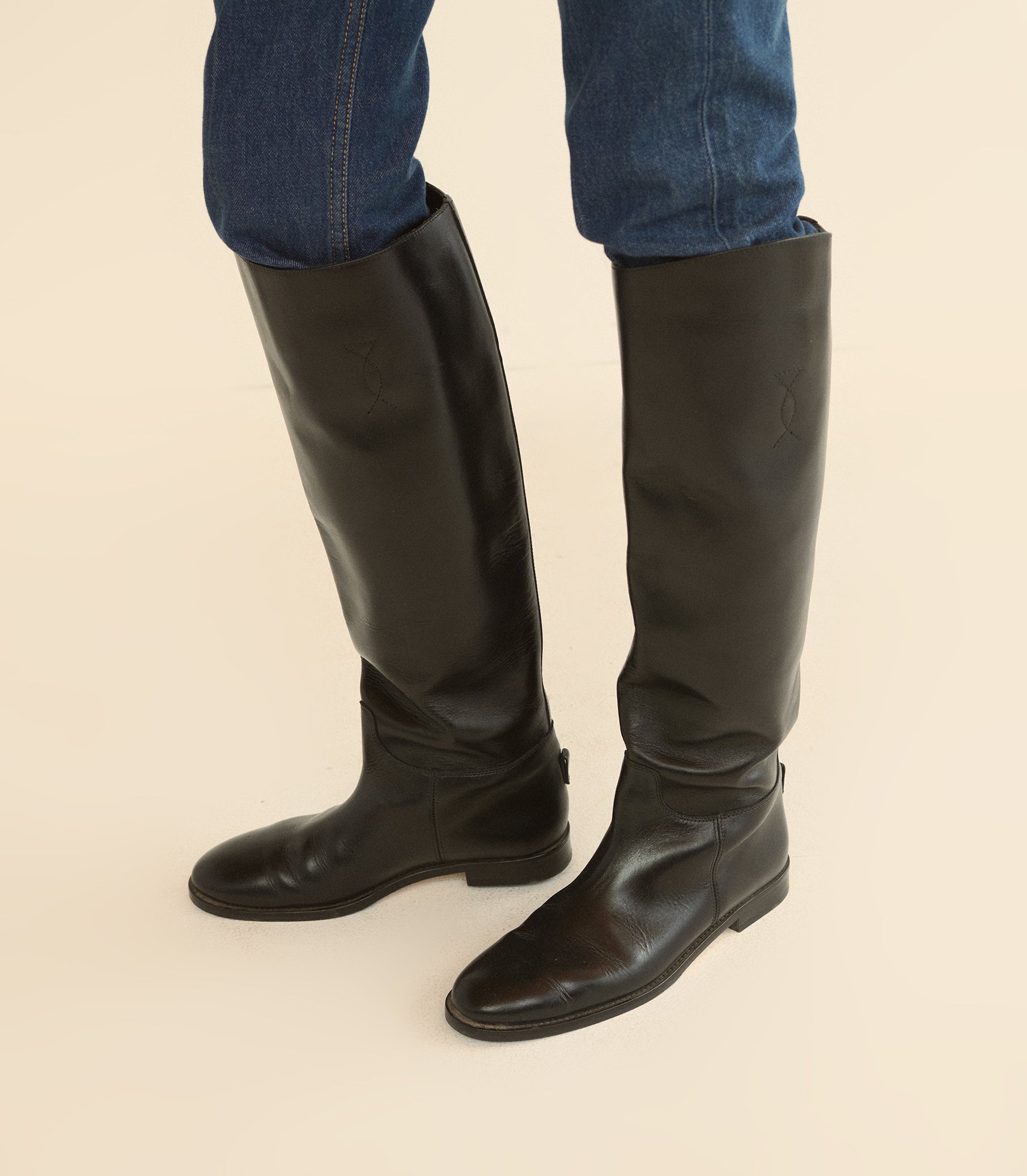 A pair of black leather knee-high riding boots worn with blue jeans, shown from the calves down against a solid light beige background.