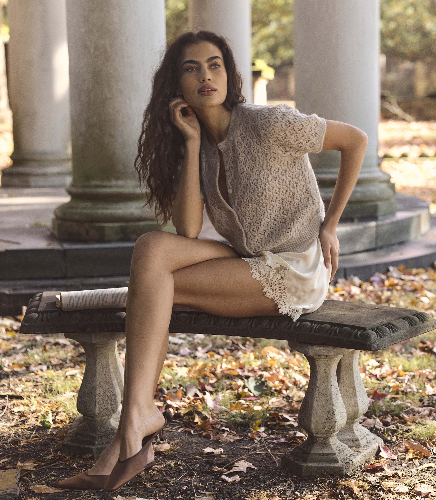 Woman wearing a beige short sleeve cardigan sitting on a stone bench in a park with columns in the background 1