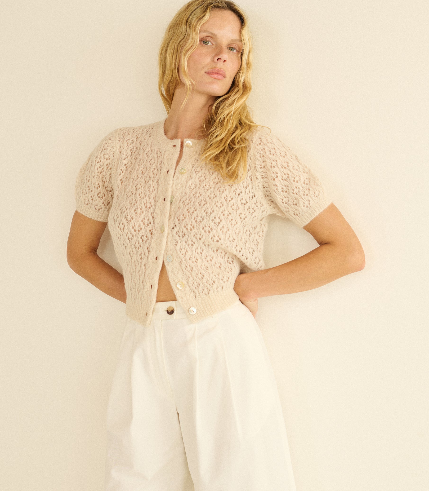 Woman wearing a beige knitted cardigan over a white top and pants on a plain background