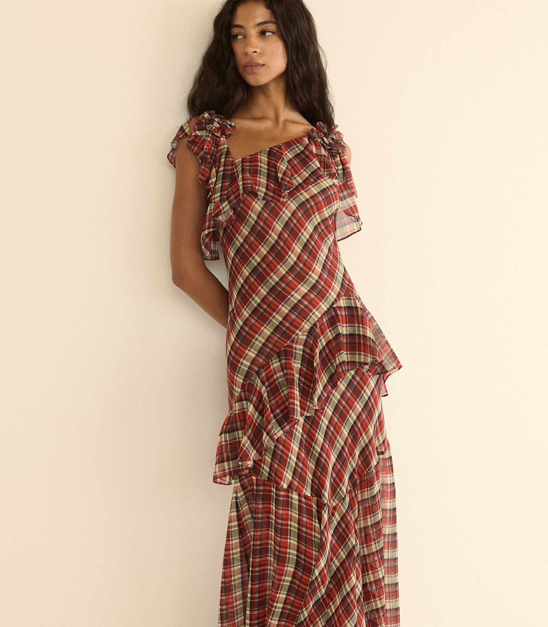 A woman wearing a red and cream plaid ruffled dress, posing against a plain beige wall.