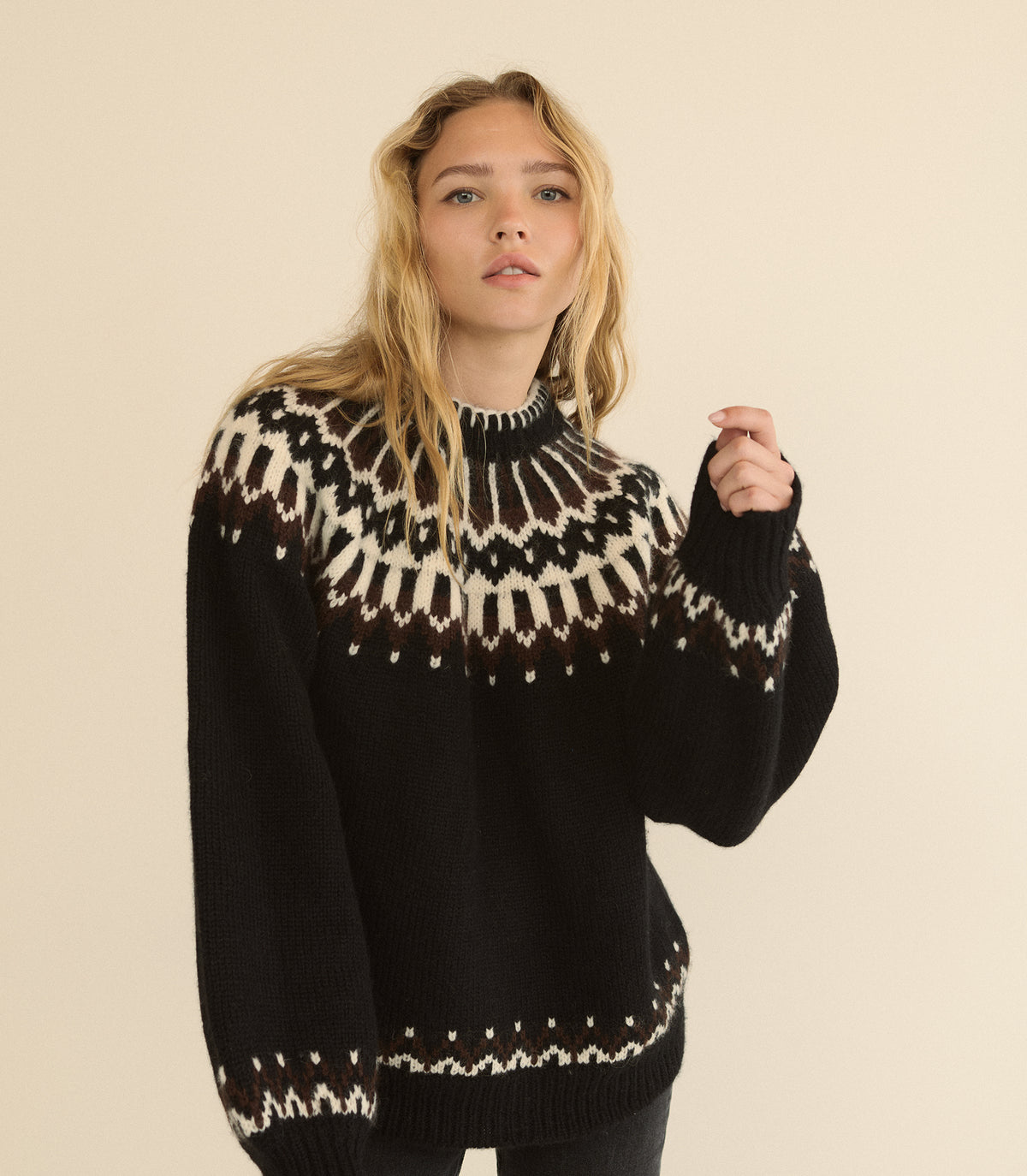 A woman wearing a black knit sweater with a cream and brown Fair Isle yoke pattern, photographed against a solid beige studio background. 1
