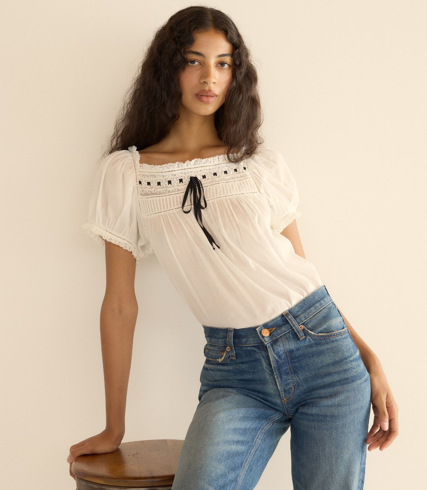 Woman wearing a white blouse with lace details and blue jeans against a beige wall.