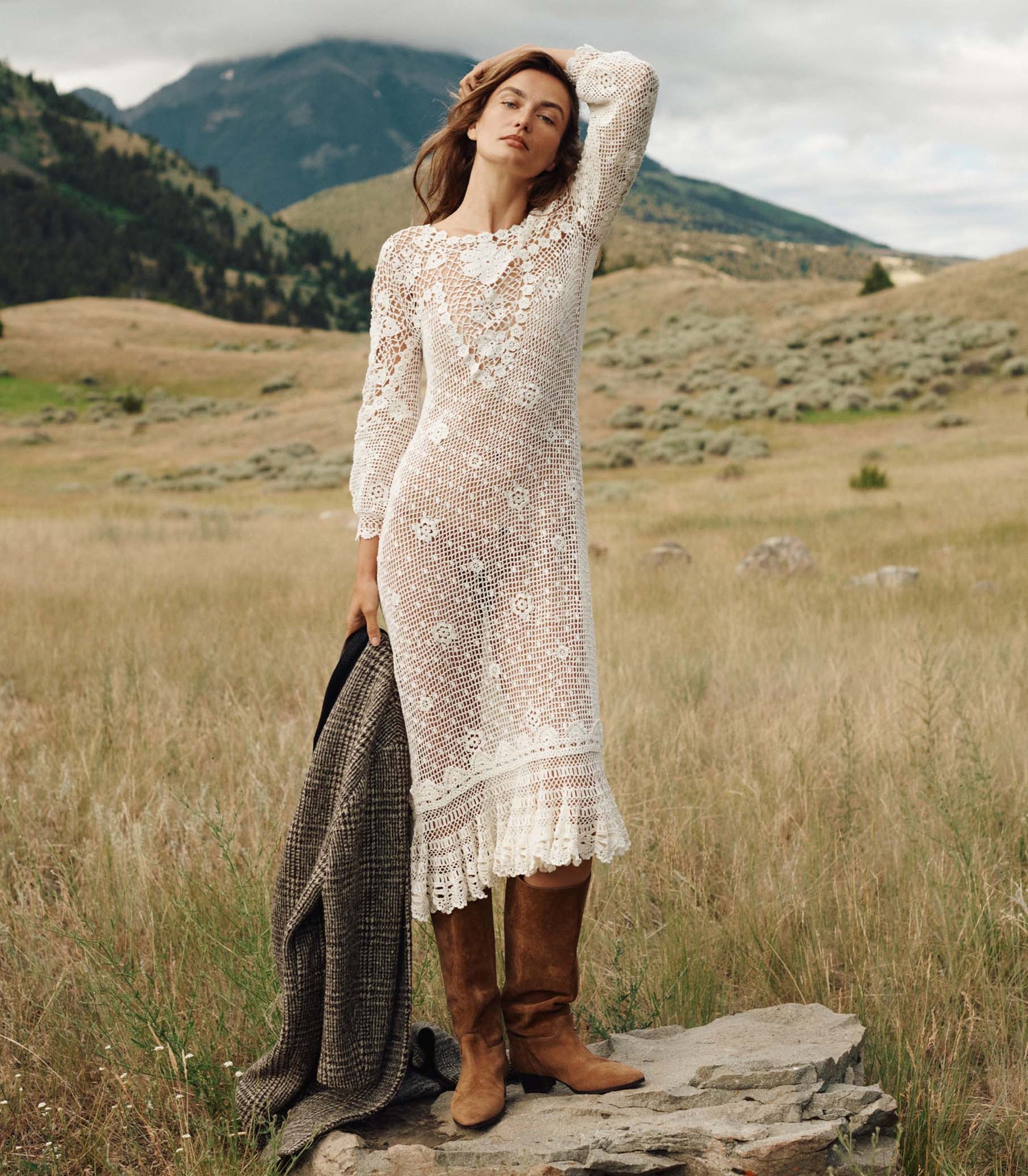 Woman in a white lace dress and brown boots standing in a field with mountains in the background
