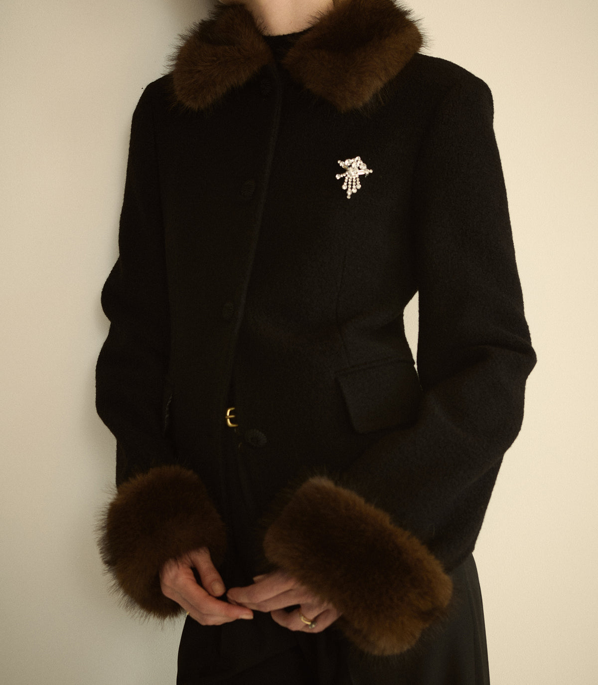 A person wearing a black coat with dark brown fur trim at the collar and cuffs, featuring a small white embroidered bird detail on the left chest.