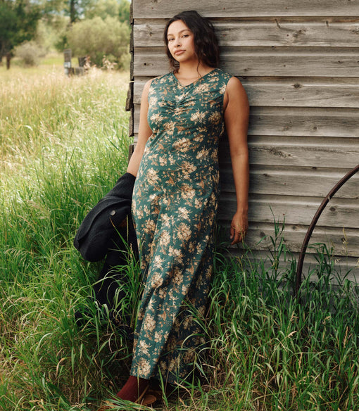 Woman in a floral dress standing in a grassy field next to a wooden building.