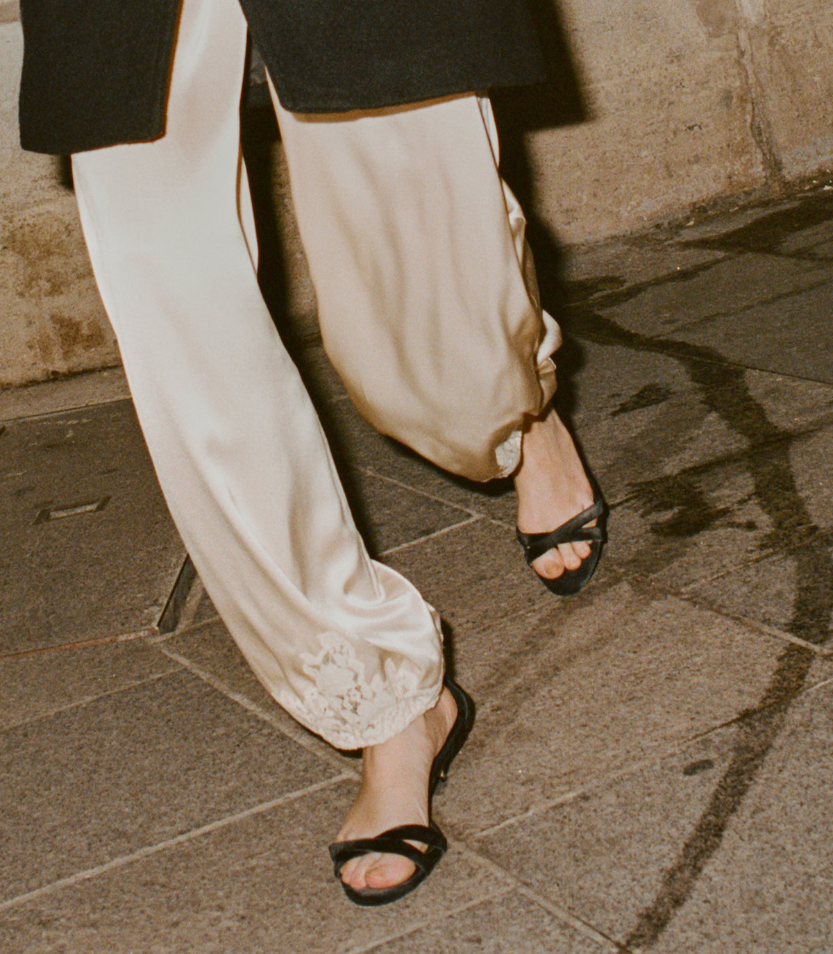 A close-up lifestyle shot of a person walking on a stone pavement, wearing cream-colored satin pants with a lace hem and black slide sandals. 1
