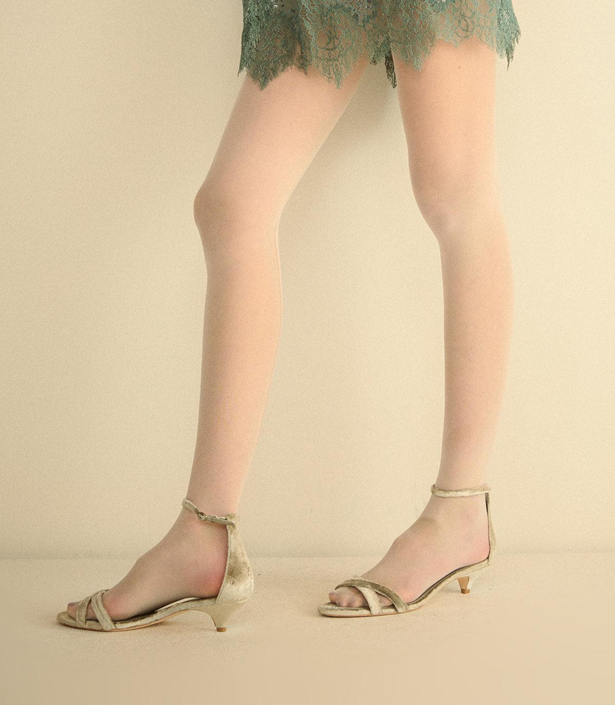 A person wearing sheer nude tights and metallic gold strappy low-heel sandals, shown from the waist down against a plain beige studio background. 1
