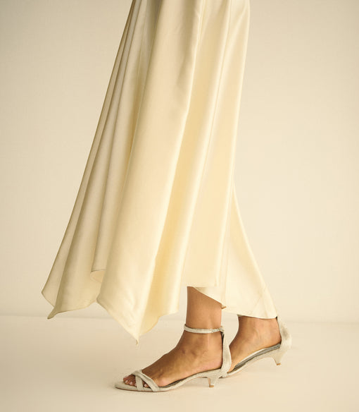 A close-up of a person's feet wearing cream satin sandals with low kitten heels and ankle straps, standing in front of a flowing cream satin skirt against a warm beige studio background.