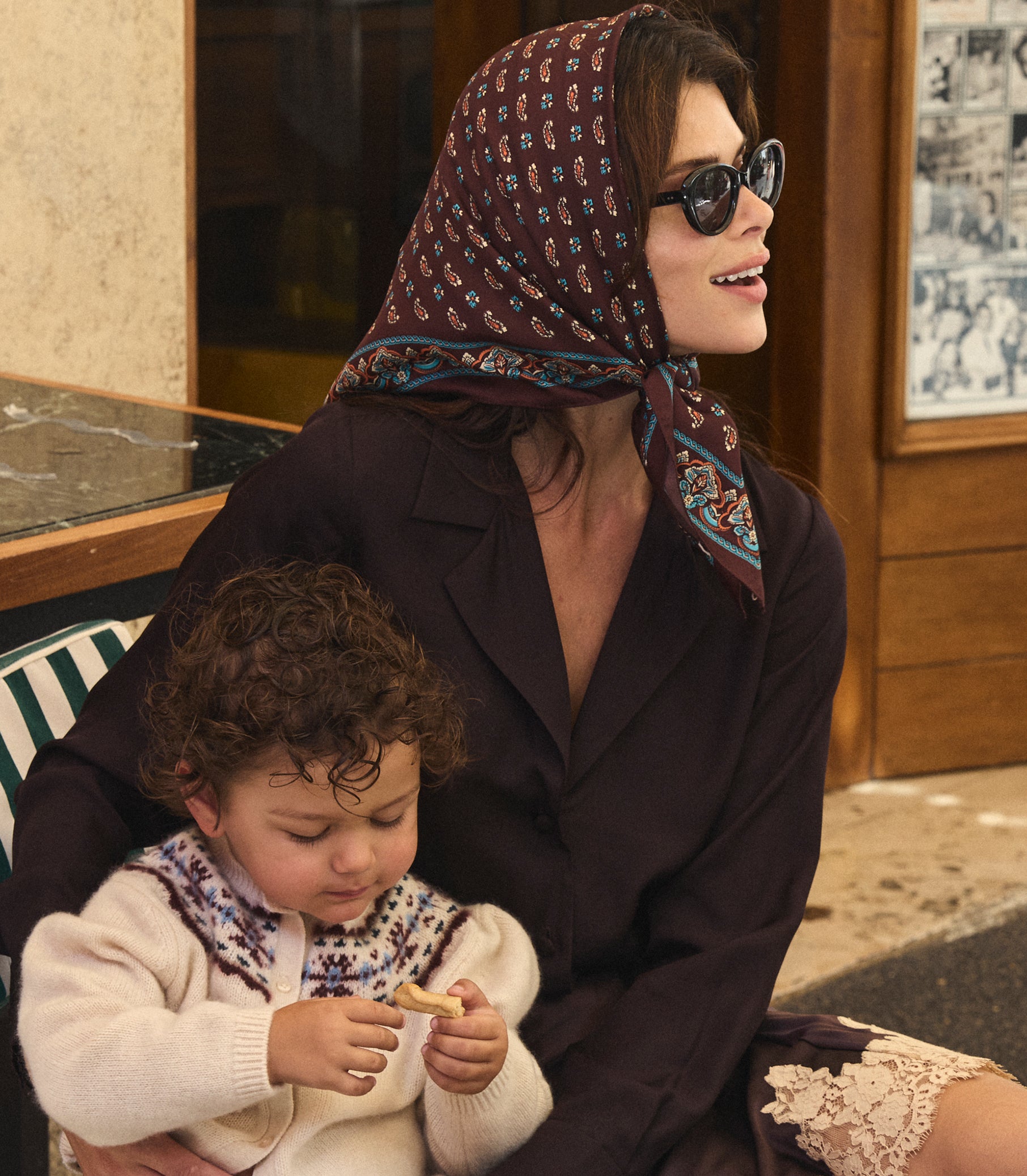 A woman wearing a dark brown patterned headscarf and sunglasses sits outdoors holding a young child. The woman's outfit includes a dark brown button up shirt and a lace-trimmed skirt, while the child wears a cream knit sweater with a patterned collar. 1
